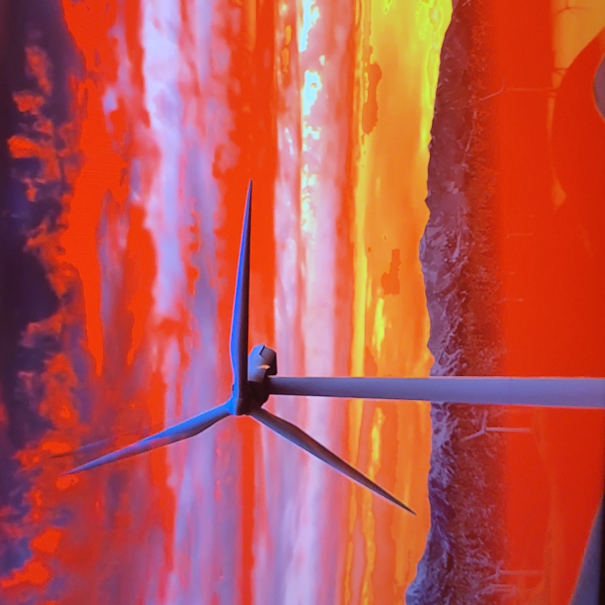 A close-up, tilted, full-frame photograph depicts a non-functional windmill with three blades. The blades are blue, and the windmill is situated against a backdrop of blended colors, primarily orange, red, and purple. The windmill appears to be stationary, and the colors in the background are blurred, giving an abstract impression. The scene's location and context within the city of Yerevan, Armenia, cannot be determined from the provided image alone.