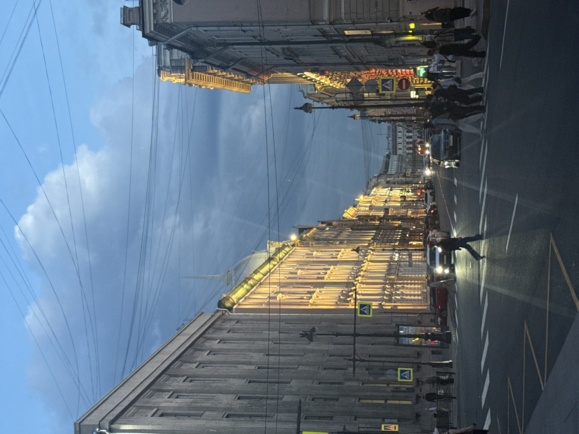 This image captures a vibrant evening street scene in Saint Petersburg, Russia, looking down a broad avenue flanked by imposing, ornate buildings. The time of day appears to be twilight or early night, as the sky shows a gradient from lighter blue with scattered clouds on the upper left to a deeper, darker blue/grey towards the right. A dense web of power lines stretches across the sky.
The grand, multi-story buildings lining both sides of the street are dramatically illuminated with warm, golden lights, highlighting their intricate architectural details, columns, and cornices. One building on the left features a distinct, slender spire rising above its roofline. The street is active with both vehicular and pedestrian traffic. Numerous cars are visible on the road, their headlights and taillights casting streaks of light. A prominent detail is a person crossing the street in the immediate foreground, brightly lit by the headlights of a car, creating a strong contrast against the darker road. Other pedestrians can be seen walking on the sidewalks and gathered at a crosswalk further down the street.
Road markings, including a white-striped pedestrian crossing and lane dividers, are clearly visible on the dark asphalt, which reflects the streetlights and vehicle lights. Various traffic signs are present, including multiple yellow diamond-shaped pedestrian crossing signs with a black walking figure, and a red circular "no entry" sign with a white horizontal bar. The scene conveys the bustling atmosphere of a historic city after sunset, with the illuminated architecture providing a warm and inviting glow against the fading daylight.