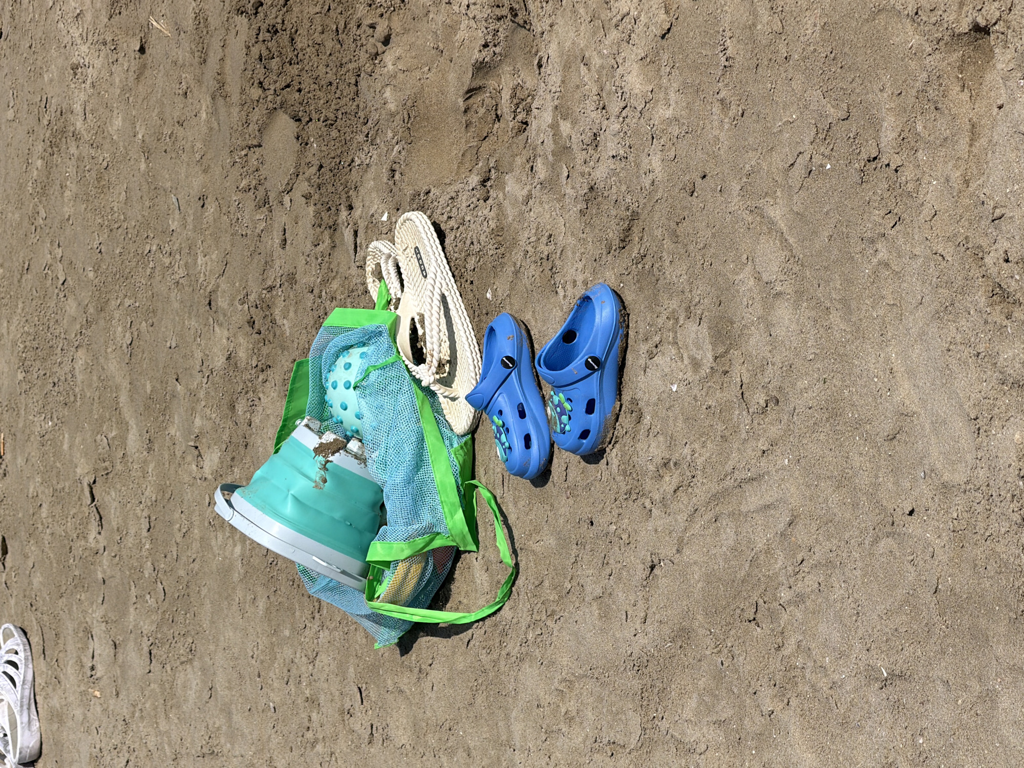 In Gatteo, Italy, on a sunny day at the beach, a collection of beach items lies on the sand. In the foreground, a pair of bright blue Crocs is arranged side-by-side, facing forward. Behind them, a pair of off-white braided flip-flops rests on the sand. A blue mesh bag with bright green trim is situated behind the flip-flops, containing a collapsible teal bucket. The bucket appears partially filled with sand and a teal ball with white polka dots. Further in the background, a white sandal can be seen lying on the sand. The environment is a sandy beach with some visible footprints and small shells scattered across the surface. The scene suggests a recent presence of people enjoying beach activities.