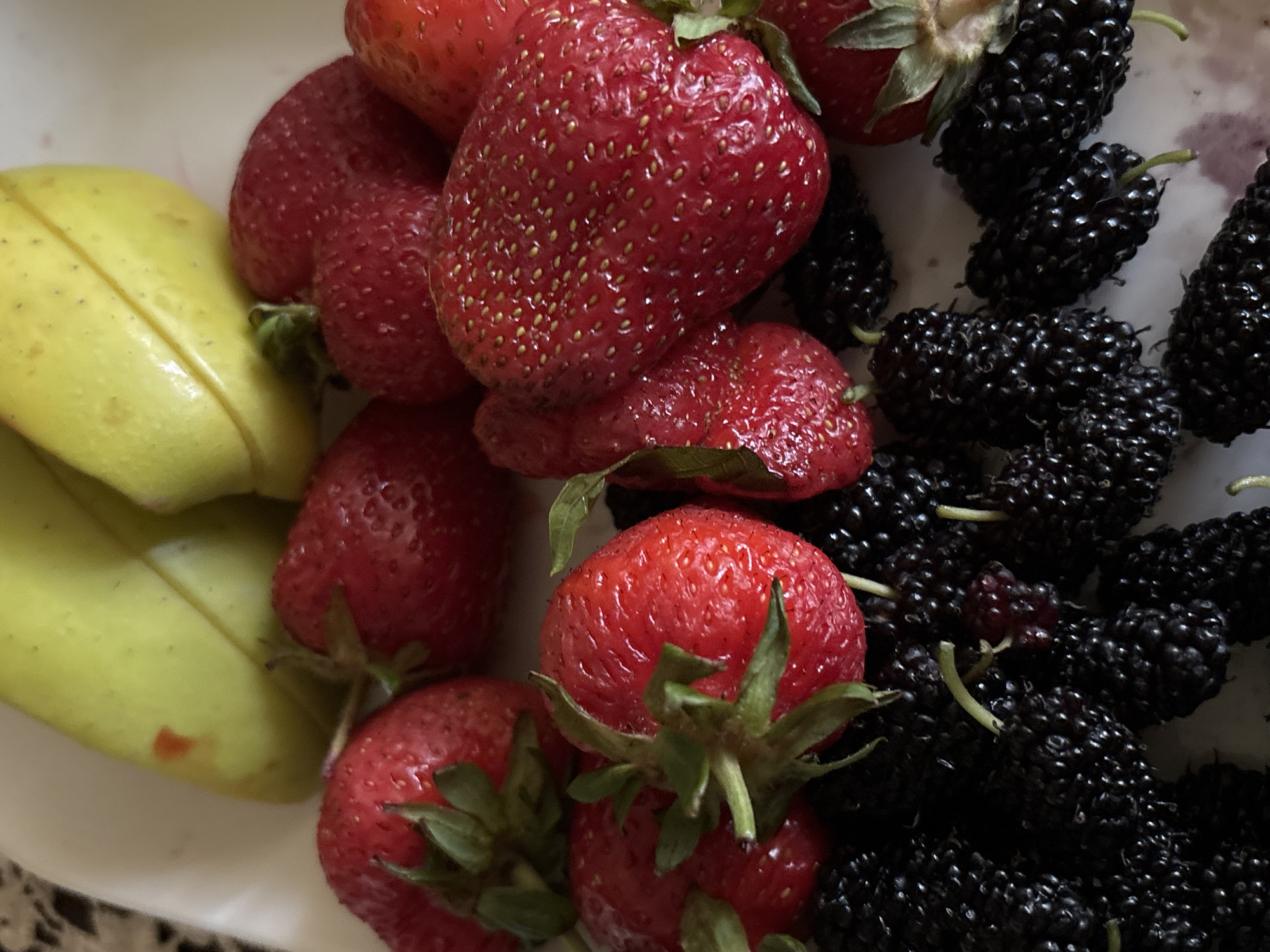 A close-up, high-angle shot presents an assortment of fresh fruits, likely arranged in a white bowl or dish, in Vagharshapat, Armenia. The dominant subjects are vibrant red strawberries and dark, almost black mulberries, with distinct yellow-green fruit slices also visible on the left.
The strawberries are plump and appear ripe, displaying their characteristic bright red color and tiny visible seeds on their surface. Many retain their green leafy caps and stems. They are clustered towards the center and right side of the frame, with some strawberries appearing larger than others.
To the right, a generous quantity of mulberries is visible. These berries are deep purple to black, showcasing their segmented texture, and several have slender, light green stems still attached. They are densely packed together, contrasting sharply in color with the bright red strawberries.
On the left side of the image, several thick, elongated slices of what appear to be yellow-green mango are stacked. These slices show a pale yellow-green color, suggesting they might be an unripe variety or a specific type of mango. They have a smooth, firm appearance with a slight gloss.
The fruits are arranged in a visually appealing composition, highlighting their natural colors and textures. The background is a clean white, likely the interior of a bowl, which serves to make the colors of the fruit stand out. There is no activity depicted, only a still life of freshly prepared fruits. No text is visible in the image.