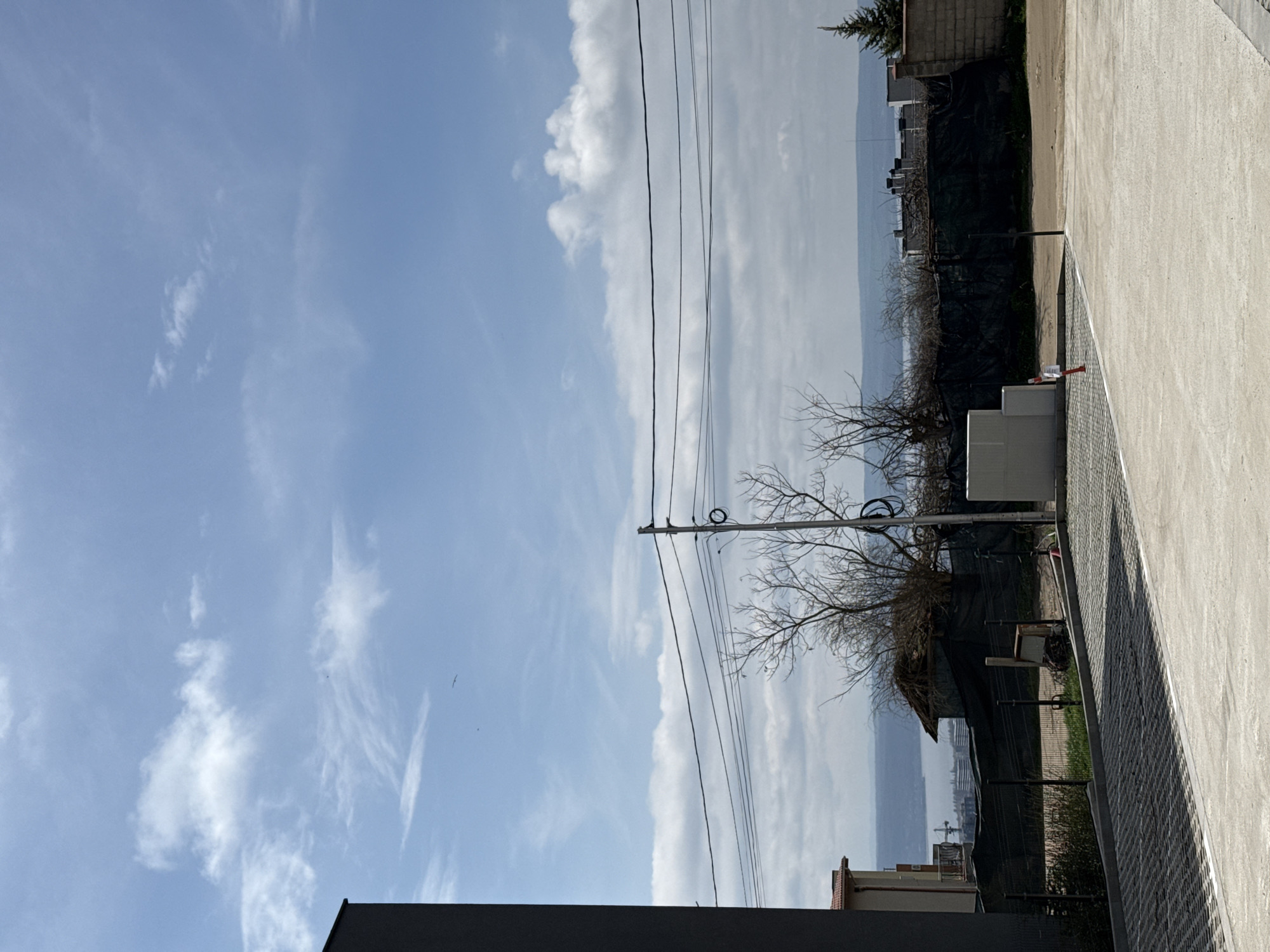 The image shows an outdoor scene under a partly cloudy blue sky. Electrical wires stretch across the sky. In the distance, a body of water is visible. The foreground features a building or structure, along with a concrete area, a fence, and some vegetation. The location appears to be in an urban or suburban area, but the image provides insufficient information to confirm it being Varna, Bulgaria.
