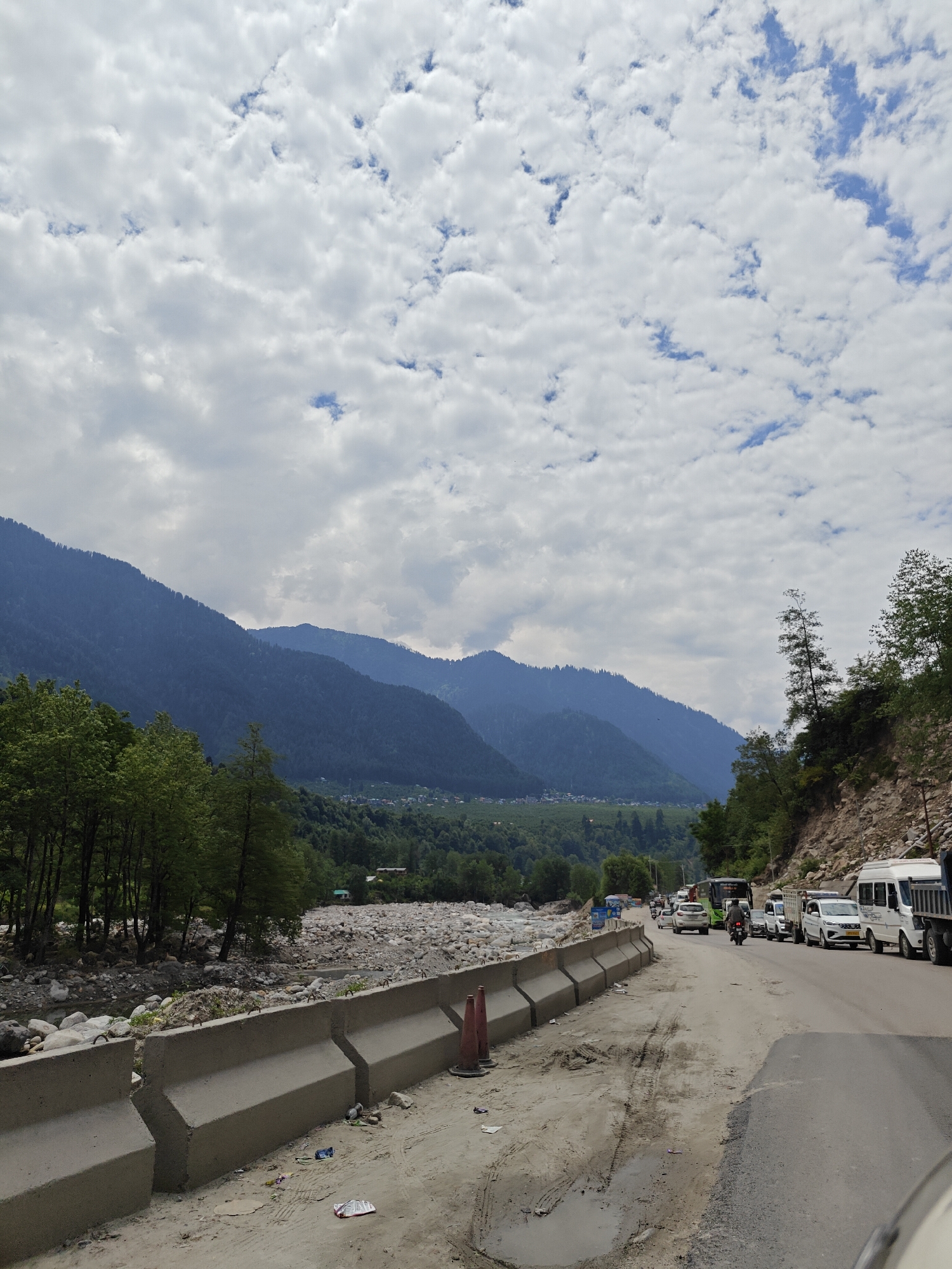 The image captures a picturesque yet busy road scene set against a dramatic mountainous backdrop, likely in Kalath, India. The sky is predominantly overcast with a mix of fluffy white and grey clouds, through which patches of blue sky are visible, suggesting an intermittent cloudy day. Towering, majestic mountains, densely forested with deep green trees, dominate the background, appearing progressively bluer and more hazy in the distance. A small village or settlement is discernible at the base of these mountains in the mid-ground.
To the left of the road, a wide, rocky riverbed stretches alongside, with some water flowing through its channels. This riverbed is flanked by trees with lighter green foliage, contrasting with the darker forests on the mountain slopes. The immediate foreground features an unpaved shoulder of dirt and gravel, separated from the river by a series of sturdy grey concrete traffic barriers. Scattered litter, including plastic bags and wrappers, lies on this shoulder, alongside a couple of orange traffic cones.
The paved road on the right is experiencing a noticeable traffic jam. A variety of vehicles are either stopped or moving slowly, bumper-to-bumper. These include several white cars (such as a Maruti Suzuki Swift, a white van, and an Eeco), a large white truck, a green bus, and a motorcycle rider navigating through the congestion. On the far right, a steep, rocky embankment, sparsely covered in vegetation, rises alongside the road.
The scene is illuminated by diffused daylight, indicative of an overcast day. No specific people are clearly visible, and there is no discernible text on any objects or signs. The overall impression is one of a popular mountain route, blending natural beauty with the everyday hustle of travel and transportation.
