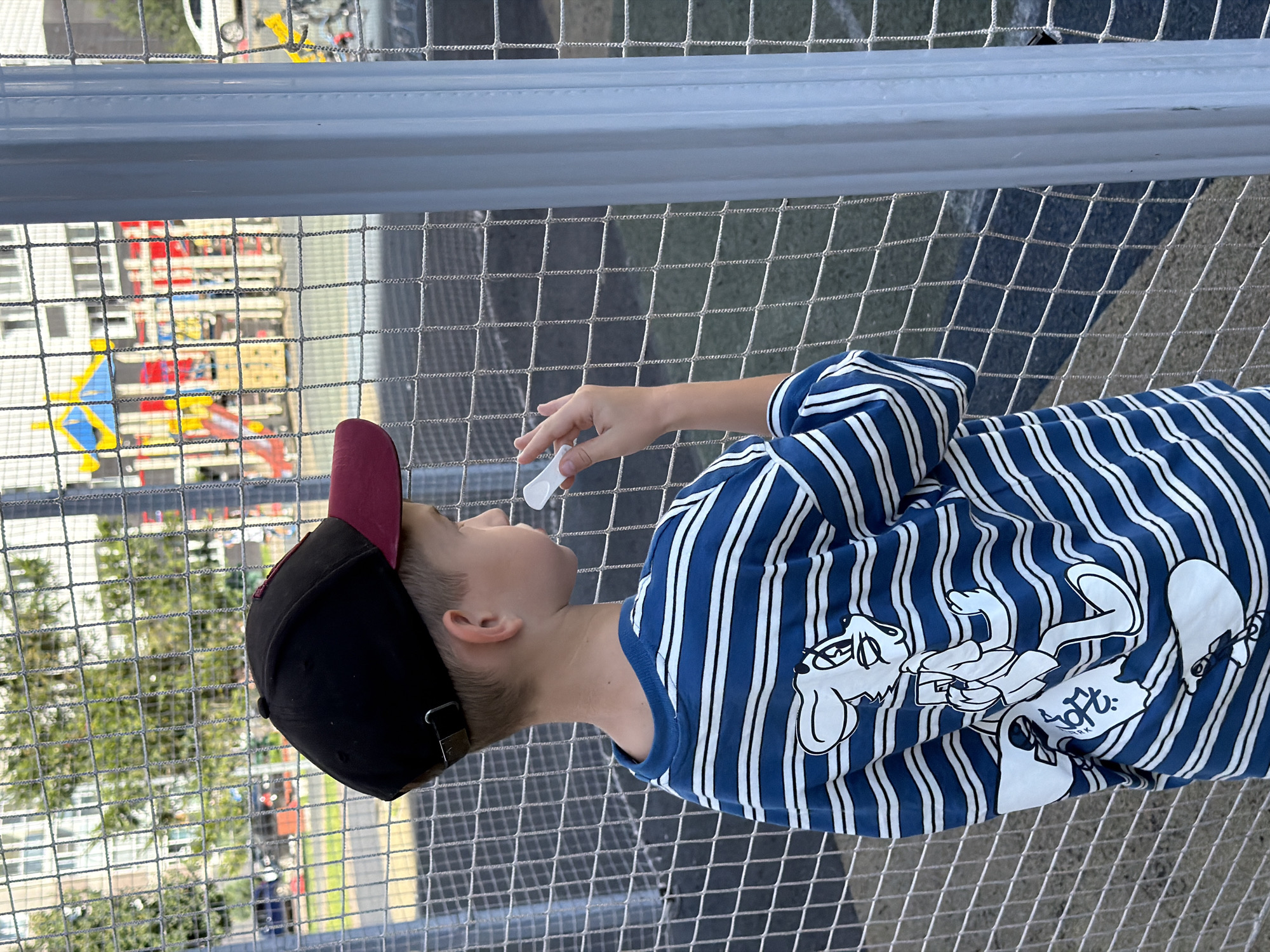 An outdoor medium shot captures a young boy, seen from behind and to the side, standing near a chain-link fence. He is wearing a black and maroon baseball cap, a blue and white horizontally striped long-sleeved shirt with a cartoon character graphic on the back, and a black baseball cap with a maroon brim. The boy is holding a white object, possibly chalk, to his mouth and looking towards a playground in the background. The playground features colorful slides and structures, and is situated behind the fence. In the far background, a building with windows and trees can be seen. The overall setting appears to be an urban area, likely a park or schoolyard. While the location is specified as Kazan, Russia, this cannot be visually confirmed from the image alone.