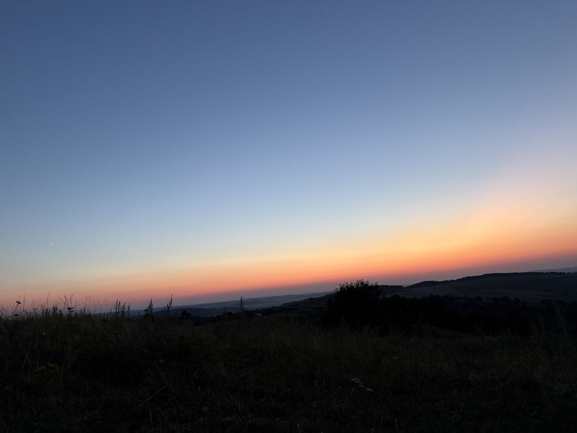 This image depicts a serene, natural landscape at dusk in an unknown location. The sky transitions from a deep blue at the top to a warm gradient of orange and pink near the horizon, indicating the setting sun. The foreground is dominated by dark silhouettes of tall grass and wildflowers, suggesting a meadow or a field. In the middle ground, rolling hills are visible, also in shadow, with some sparse trees or bushes dotting the landscape. The overall impression is one of peacefulness and natural beauty, with no human presence or man-made structures immediately apparent.
