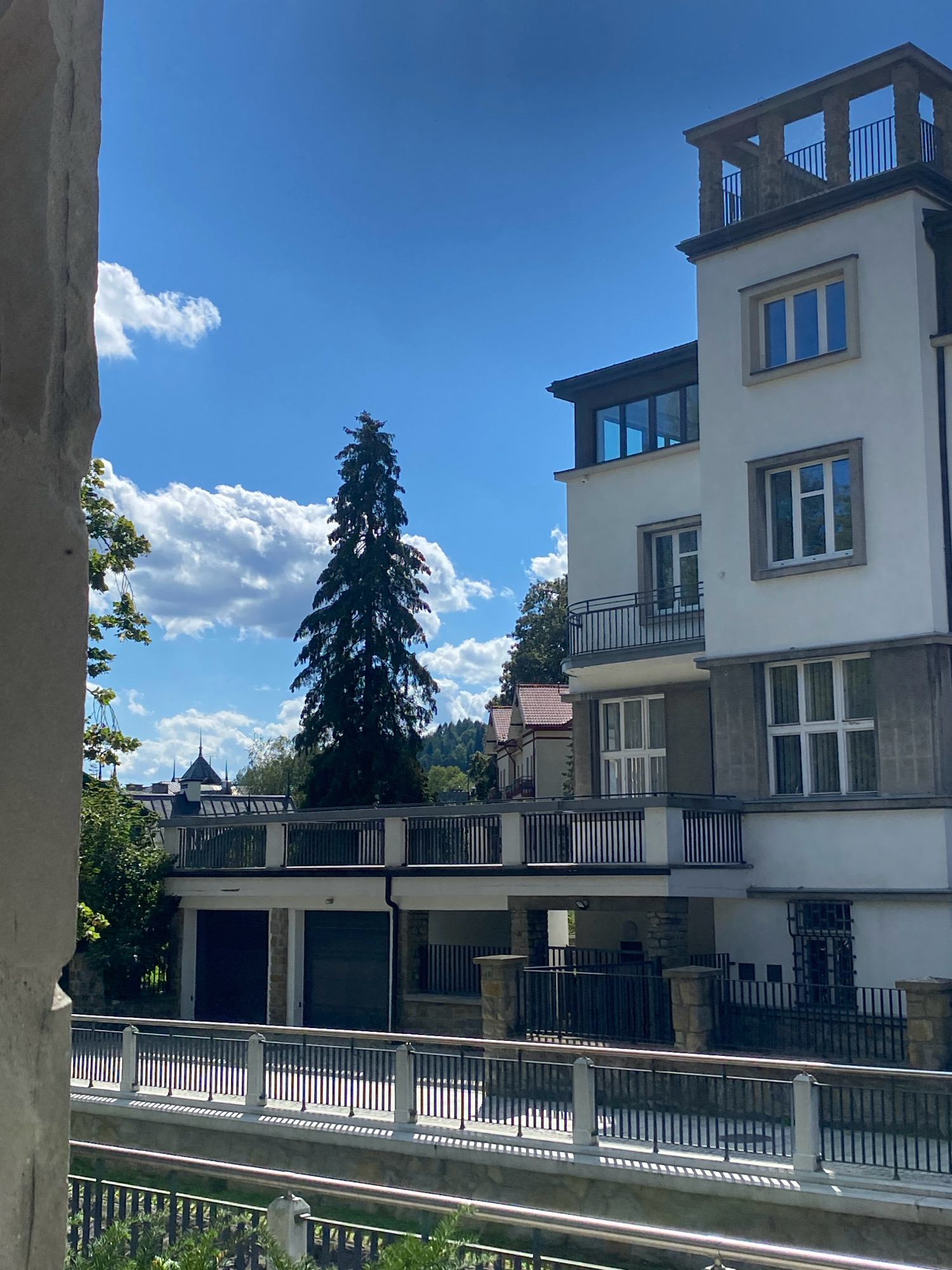 The image shows a modern white building with balconies and large windows, located in the city of Krynica-Zdrój, Poland. In the foreground, there is a railing and greenery. A tall, dark green coniferous tree stands to the left of the building, partially obscuring it. The sky is blue with scattered white clouds. In the background, other buildings with red roofs and a treed hillside are visible. The architectural style suggests the building is relatively new, possibly a hotel or apartment complex. The overall scene is a bright, sunny day.