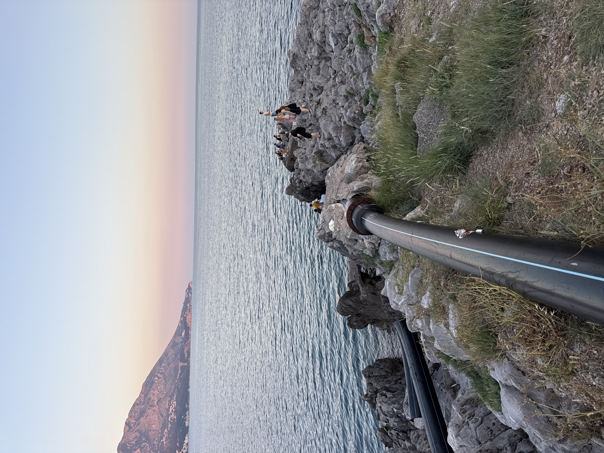 In Boreti, Montenegro, a wide shot captures a rocky coastline at dusk. The sea, with a gentle ripple, fills the lower half of the frame, transitioning to a sky painted with soft hues of pink and blue. On the left, two large black pipes, possibly for water or sewage, lie partially embedded in the rocks, stretching towards the water. A prominent pipe, thick and black with a thin blue stripe, runs diagonally across the foreground from the bottom right towards the sea. In the mid-ground, a cluster of large, grey rocks juts out into the water, serving as a gathering point for approximately eight individuals. Some are seated, while others stand, appearing to be engaged in conversation or relaxation. One person stands slightly apart on the highest point of the rocks, observing the scene. The rocky terrain is interspersed with patches of dry grass and vegetation. In the distance, a mountainous silhouette frames the horizon, its slopes dotted with what appear to be buildings, suggesting the presence of a town or city. The overall atmosphere is serene, marked by the gentle light of twilight.
