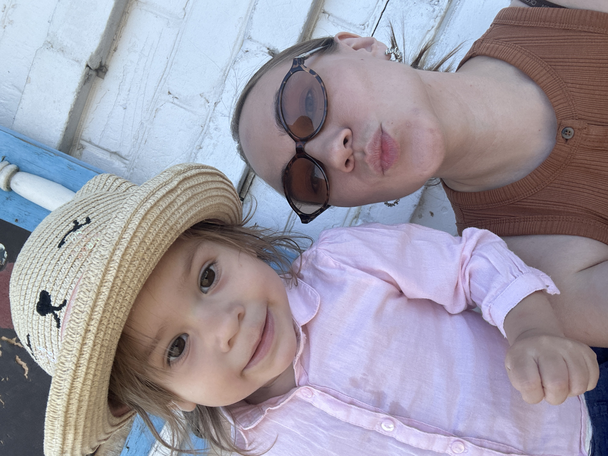 A selfie shows a woman and a young child. The woman wears sunglasses and a brown top. The child wears a straw hat decorated with cartoon bears and a pink button-down shirt. They appear to be on an outdoor bench or porch in front of a white painted wall. It is not possible to determine the city of Chaikovsky, Russia based on this image alone.