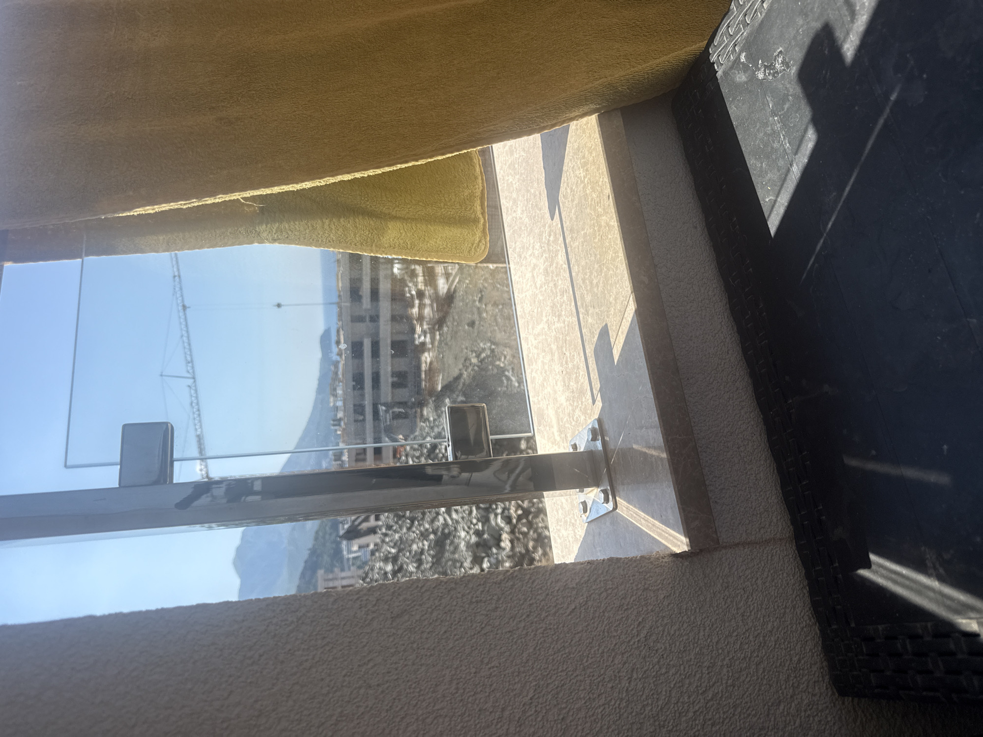 An outdoor scene is depicted through a glass railing, suggesting a balcony or patio in Boreti, Montenegro. The view looks out over a construction site with a crane visible in the distance, and a body of water can be seen beyond. Mountains are present in the background. A yellow curtain hangs to the right of the frame, partially obscuring the view. The foreground shows a black surface with a woven texture, possibly furniture, under sunlight.