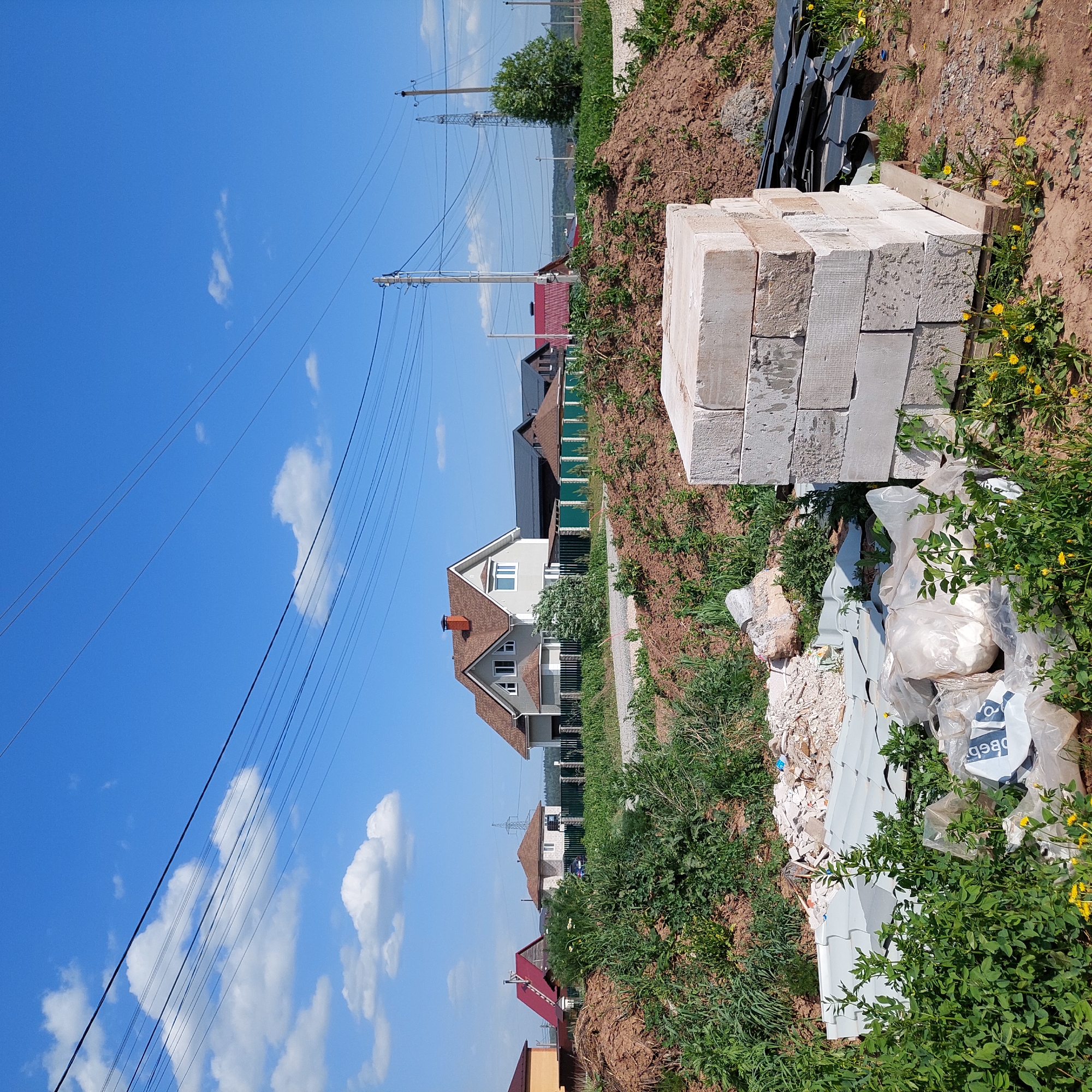The image shows a construction site in Bol'shaya Mos', Russia. A stack of concrete blocks is visible in the foreground, along with debris like scraps of metal and plastic, and overgrown vegetation. Beyond the construction site, houses with red and brown roofs, and a light-colored house with a brown roof can be seen. Overhead power lines stretch across a blue sky with scattered clouds.