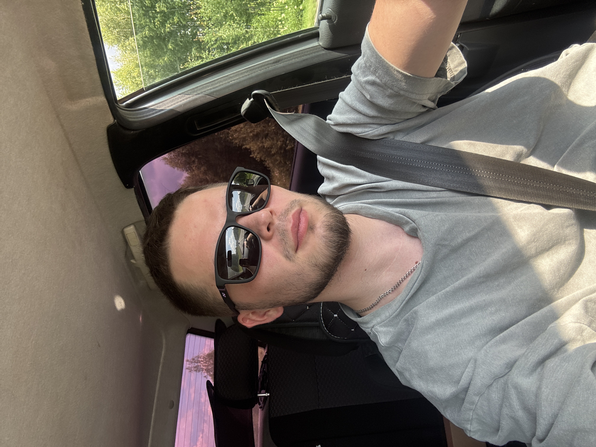 A man with short brown hair and a beard is driving a car, reflected in his sunglasses is a road and other cars. He is wearing a grey t-shirt and a seatbelt. The car interior is visible, along with trees and sky outside the windows. The scene appears to be in Marchugi, Russia, based on the information provided.