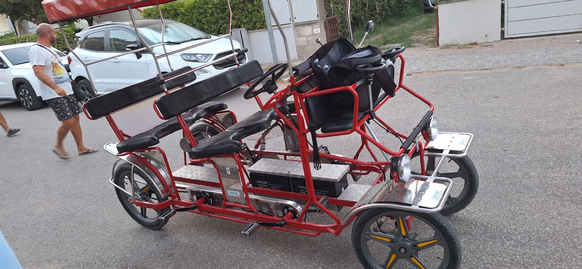 A red four-wheeled surrey bicycle with two rows of black seats and a red canopy is parked on an asphalt road in Ravenna, Italy. A man wearing a white t-shirt, patterned shorts, and flip-flops is walking past the bicycle on the left side of the image. A white SUV is parked behind the bicycle, and a white house with a brown fence is visible in the background. The surrey has two steering wheels and what appears to be a bag on the front passenger seat. The road surface is a mix of asphalt and pebbles. The image is taken from a slightly elevated angle, looking down at the surrey.