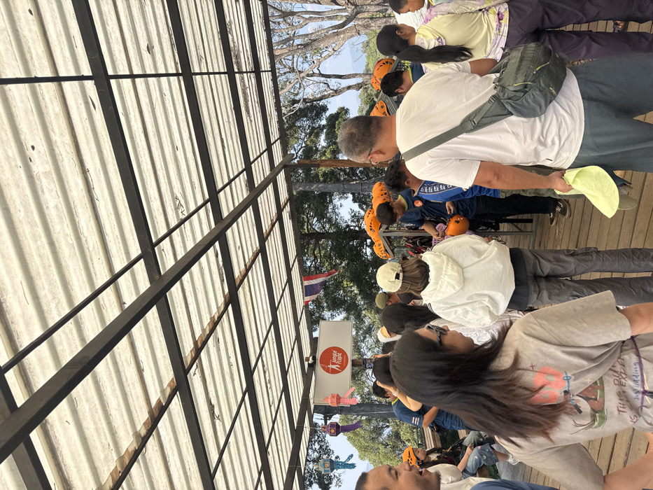 A group of people stands outdoors, some wearing orange helmets. One man wears a white shirt and gray pants, a gray bag slung across his shoulder. A woman stands in front, wearing a gray t-shirt and glasses. A Thai flag is visible in the background, next to a sign with a red logo. An open-roofed metal structure with a corrugated ceiling covers part of the area where the group is standing. Trees and the sky are visible in the background.