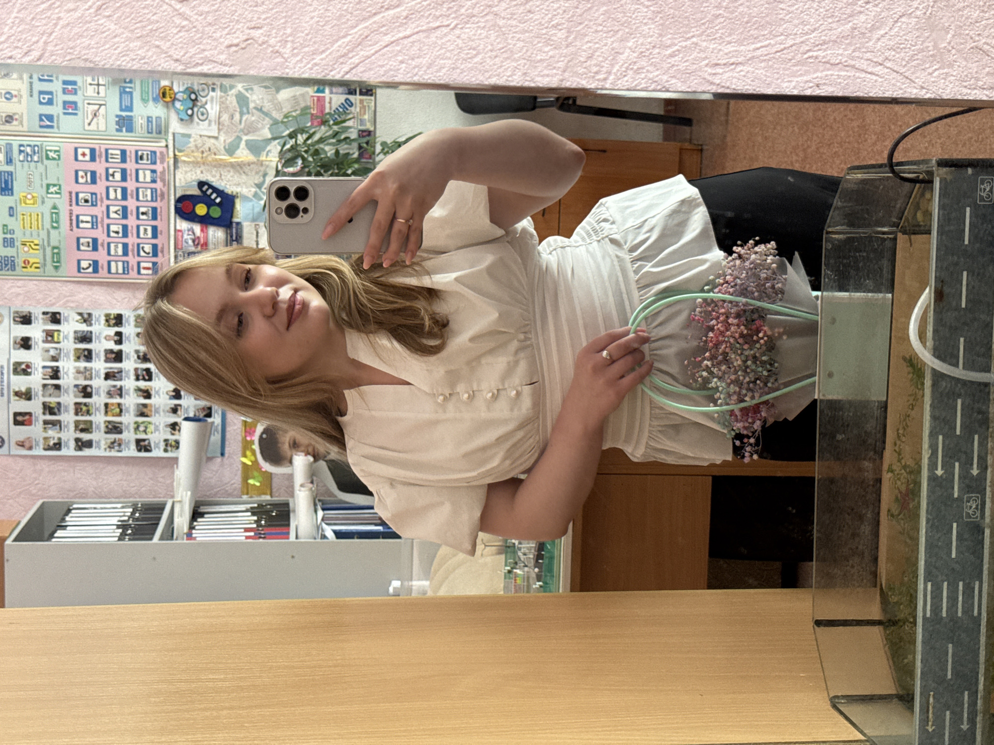A young Caucasian woman with blonde hair, wearing a white puffed-sleeve blouse and black pants, is taking a mirror selfie in what appears to be an educational or administrative setting. She is holding a small bouquet of dried flowers in a light green paper bag with a light green handle. In her right hand, she is holding a silver smartphone. She is wearing two rings, one on her right index finger and another on her left ring finger. Behind her, a bookshelf filled with binders and papers is visible. The room has a pink textured wall and a wooden desk. The floor is tiled, and a glass terrarium or aquarium with a road design is in the foreground. The overall impression is that of a casual photo taken indoors, likely in an office or classroom. The presence of educational posters on the wall suggests a school or university environment in Chaikovsky, Russia.