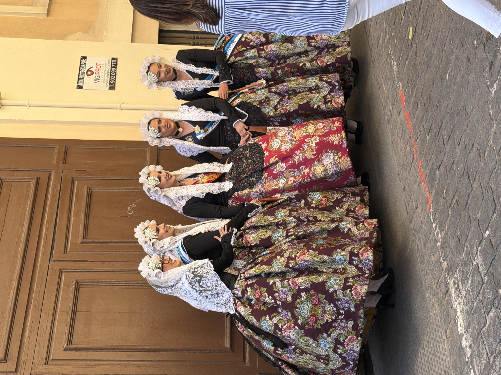 Five women are dressed in traditional Alicante clothing. Each has a white, lacy headdress, a black blouse, and a long floral print skirt. They are standing in a line on a sidewalk in front of a brown door in Alicante, Spain. A security company sign is on the wall. A person wearing white pants and a striped shirt is partially visible in the upper right corner. The sidewalk is paved with grey cobblestones.