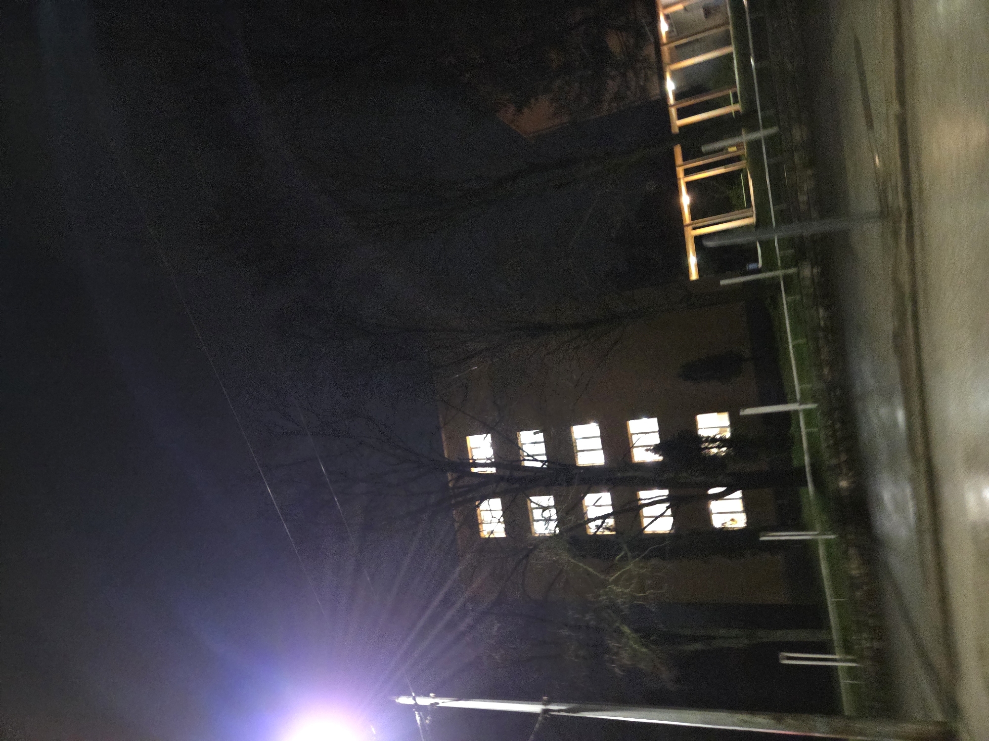 A nighttime photograph, taken at an angle, shows a building with illuminated windows in Pleven, Bulgaria. The building is a muted yellow color. The windows are arranged in a rectangular grid pattern. Bare tree branches are visible in front of the building. A bright light source, possibly a street lamp, illuminates the bottom portion of the scene, creating a glare. A structure with rectangular, illuminated elements is visible above the first building. The ground is paved and appears wet, possibly due to rain. Metal bars or railings are placed along the top. The rest of the scene is shrouded in darkness.