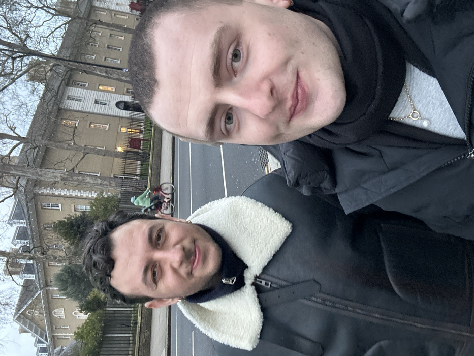 Two men pose for a selfie outdoors, with overcast lighting. The taller man on the top has light skin, short hair, and wears a black scarf and jacket. He sports a necklace. The other man has darker hair and complexion, wearing a black jacket with a large white collar. In the background, an urban street scene includes buildings, trees, a black fence, and a cyclist riding on the road.