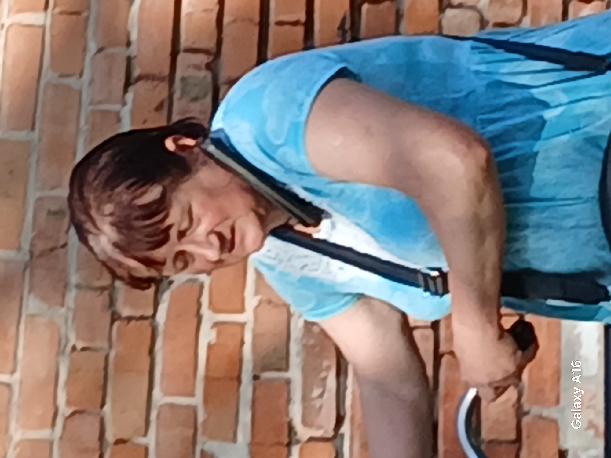 A person is captured in a close-up shot, their body angled sideways. They are wearing a light blue t-shirt with a subtle pattern. A black harness is visible over their shoulders and chest, with dark straps secured around their waist. Their hair is dark brown, styled with bangs, and their head is tilted downwards, obscuring their full facial expression. Their right arm is bent, and their hand is grasping a curved, metallic object. The background consists of a brick wall with an orange-brown hue, and the mortar between the bricks appears light. The lighting suggests it is daytime, possibly outdoors given the natural brick texture. A watermark "Galaxy A16" is visible on the right side of the image. The image appears to be taken in Ustyvytsia, Ukraine.