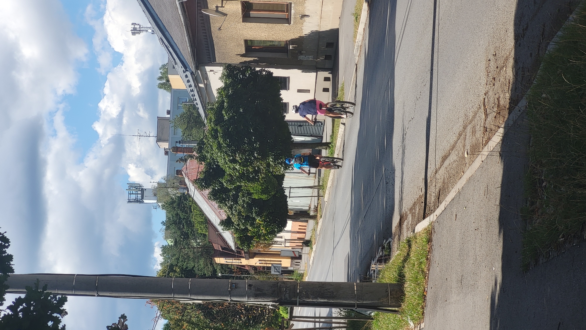 In Prešov, Slovakia, two individuals are cycling down a paved street lined with trees and buildings under a partly cloudy sky. The person in front is wearing a blue shirt and dark shorts, with a blue helmet. The person behind is wearing a pink shirt and shorts, with a white helmet. The street features a curb and grass on either side. In the background, a tall communications tower is visible above residential buildings. The scene appears to be a normal day in a residential or mixed-use urban area.