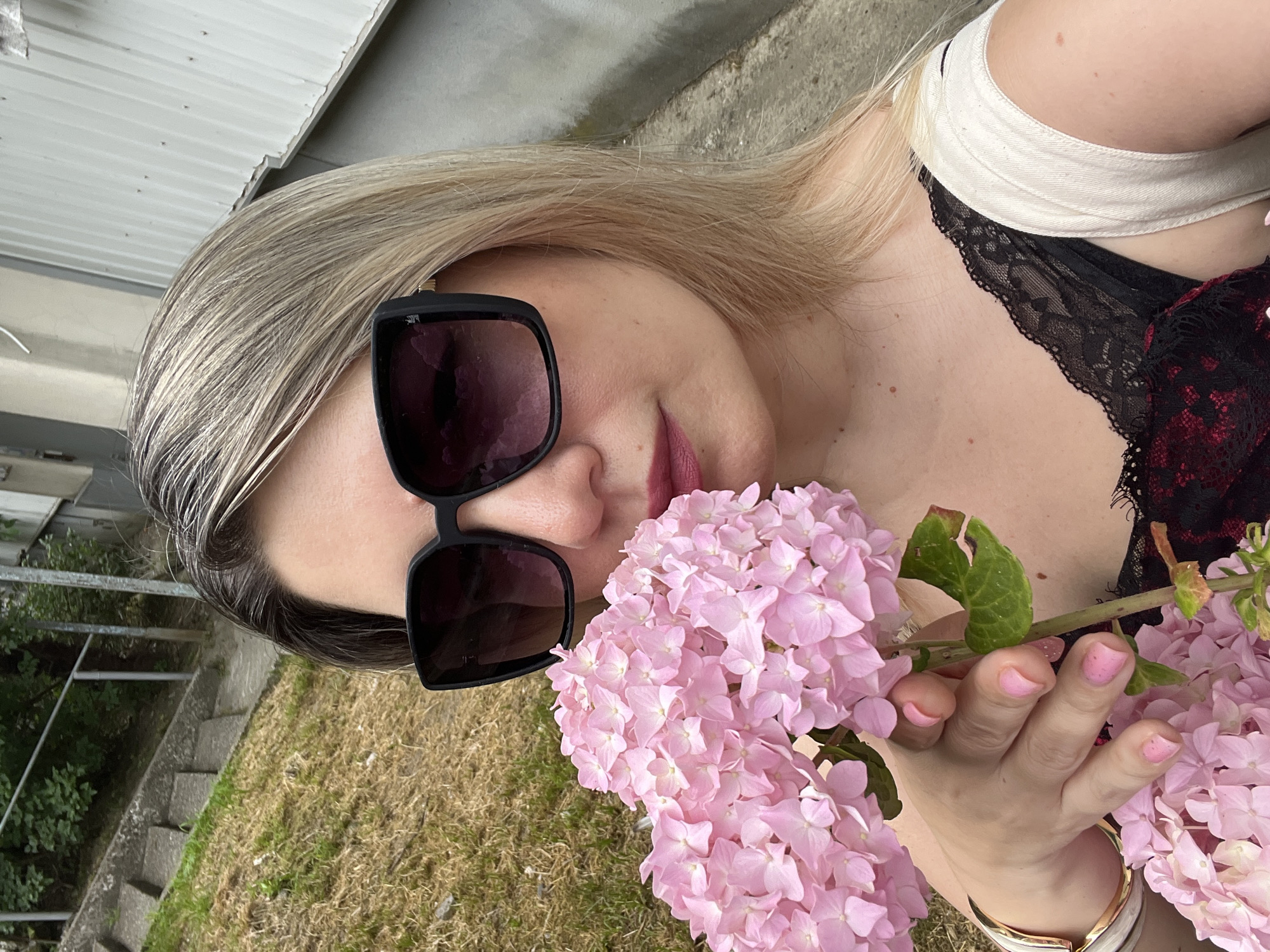 A woman with blonde hair and sunglasses holds a bunch of pink hydrangeas near her face. She is wearing a black lace camisole with a white strap. The background shows a grassy slope with concrete stairs and a metal railing. A concrete wall or building is visible to the right. The location is specified as Sochi, Russia, though no specific landmarks confirm this.