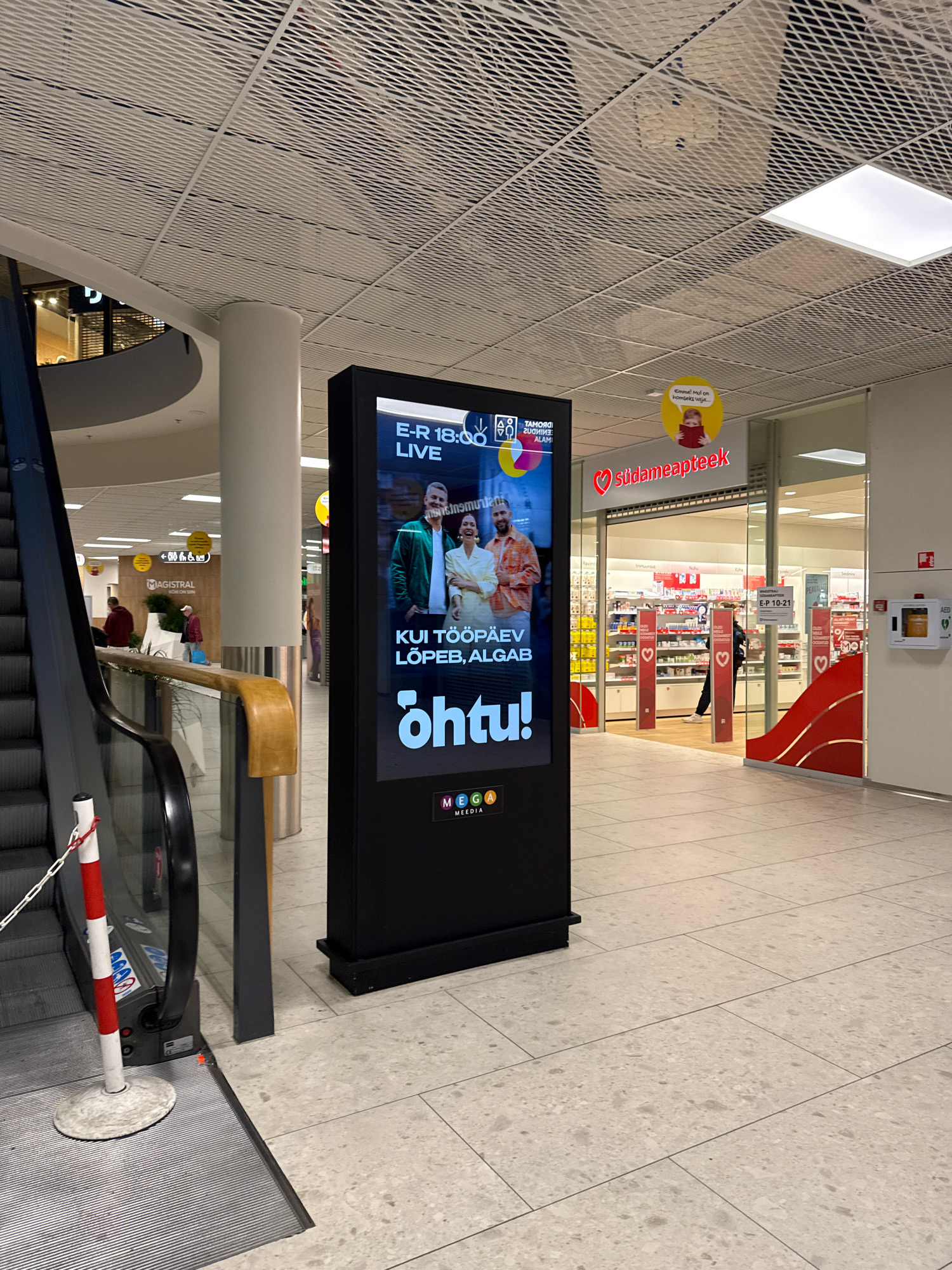 The image depicts an interior scene, likely a shopping mall, in Tallinn, Estonia. In the foreground, a black digital advertisement screen displays content related to a live broadcast, with text in Estonian. The screen shows two people, a man and a woman, smiling. Below their image, text reads "KUI TÖÖPÄEV LÕPEB, ALGAB Õhtu!" which translates to "When the workday ends, evening begins!". At the bottom of the screen, a logo with the letters "MEG" and "GA" in colorful blocks is visible.
To the left of the screen is a large escalator, with its metallic steps and handrail visible. A red and white striped pole with a chain is positioned at the base of the escalator.
In the background, there is a retail store with the name "SÜDAMEAPTEEKK" prominently displayed on its facade. The store appears to be a pharmacy, with shelves stocked with products visible through the glass doors. There are also signage and displays outside the store. Several people are seen walking in the background, some entering or exiting the pharmacy.
The environment features a modern, tiled floor and a suspended ceiling with a patterned design. Natural light seems to be supplemented by artificial lighting. The overall impression is a clean, well-lit commercial space.