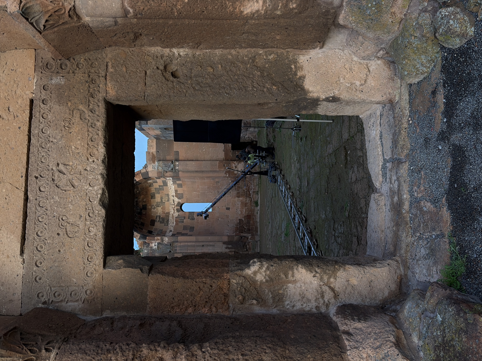 The image shows a view through a stone window or opening in what appears to be an ancient structure in Anipemza, Armenia. The opening is framed by intricately carved stone on the left side, while the stones on the right exhibit a rougher texture. Through the opening, one can see another part of the structure, which appears to be a church or cathedral, with a dome or arched roof visible. A telescopic crane or platform is positioned outside the opening, seemingly for maintenance, filming, or research purposes. Two people are visible operating the crane. The ground outside the opening appears to be a mix of gravel and vegetation.