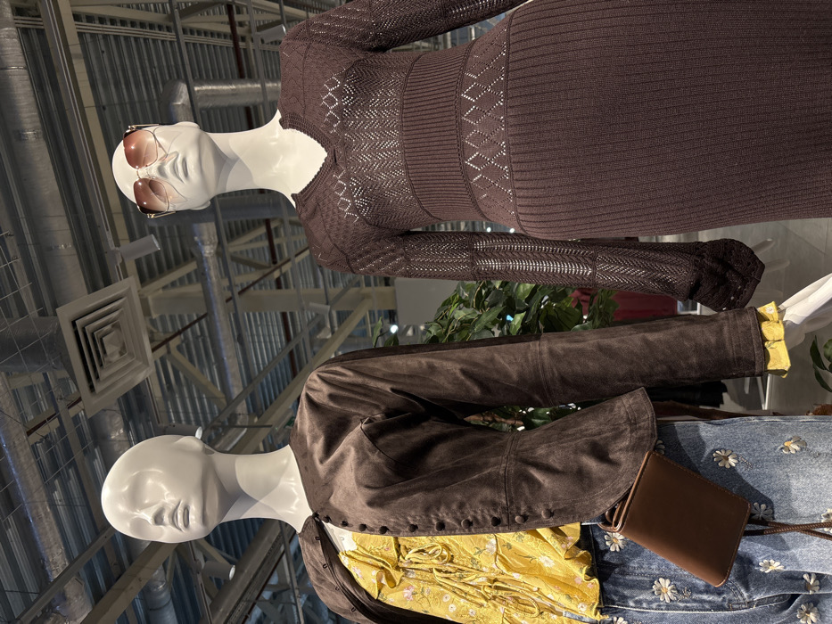 Two mannequins are on display. The top mannequin wears a brown, long-sleeved knitted dress and sunglasses. Below, another mannequin is dressed in a yellow blouse under a brown suede jacket. This outfit is paired with jeans decorated with small daisies and a brown, crossbody bag. The background shows a modern, industrial space with metal beams and ventilation systems.