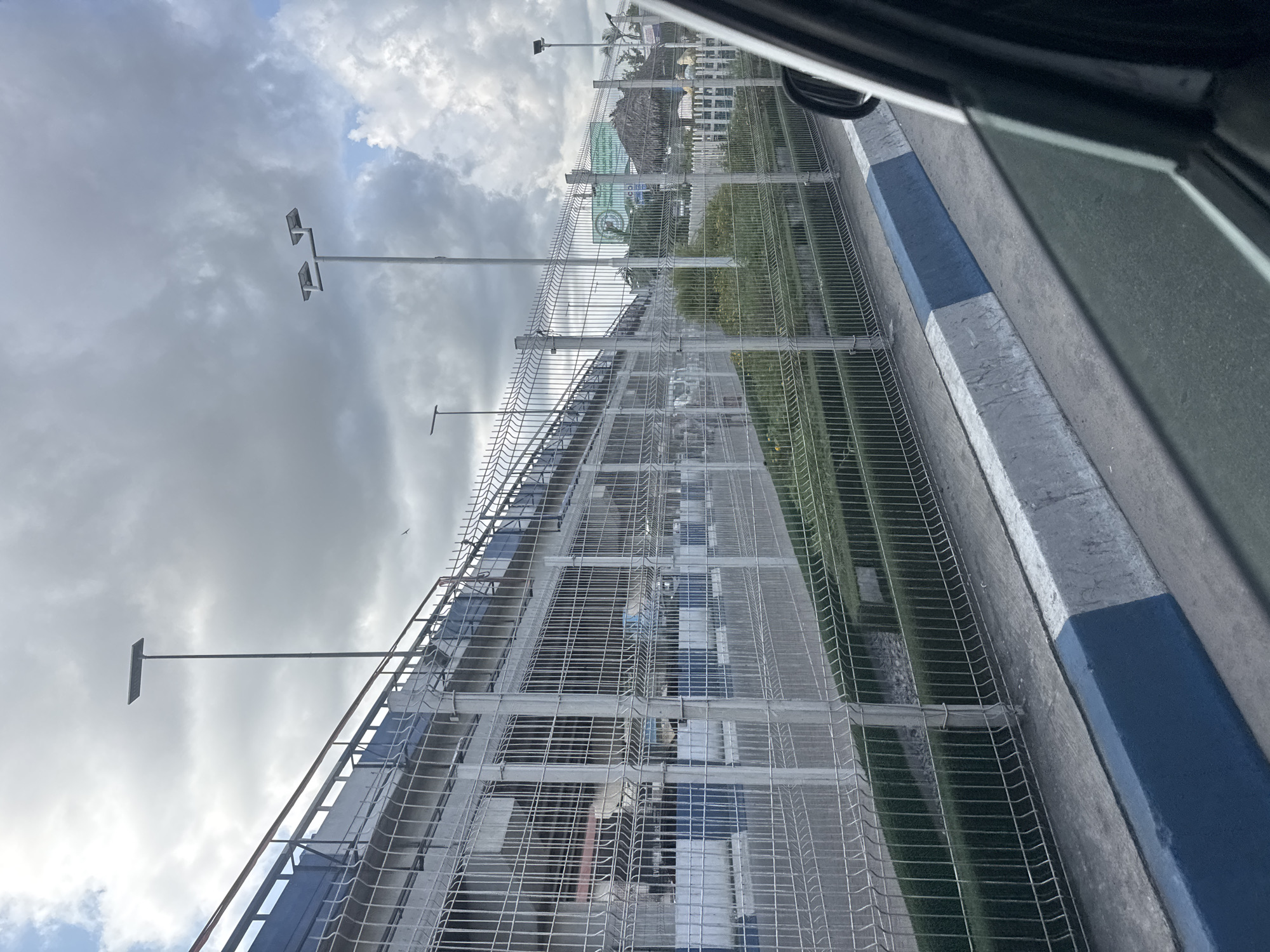 The image shows a view from inside a vehicle, looking out at a tall, wire-mesh fence topped with a ladder-like structure running parallel to a concrete curb painted with alternating blue and white sections. Beyond the fence and curb, there is grass and some landscaping, along with building structures, including one with a thatched roof. In the background, light poles extend upward towards a cloudy sky. Based on the provided information, the location is Port Harcourt, Nigeria.