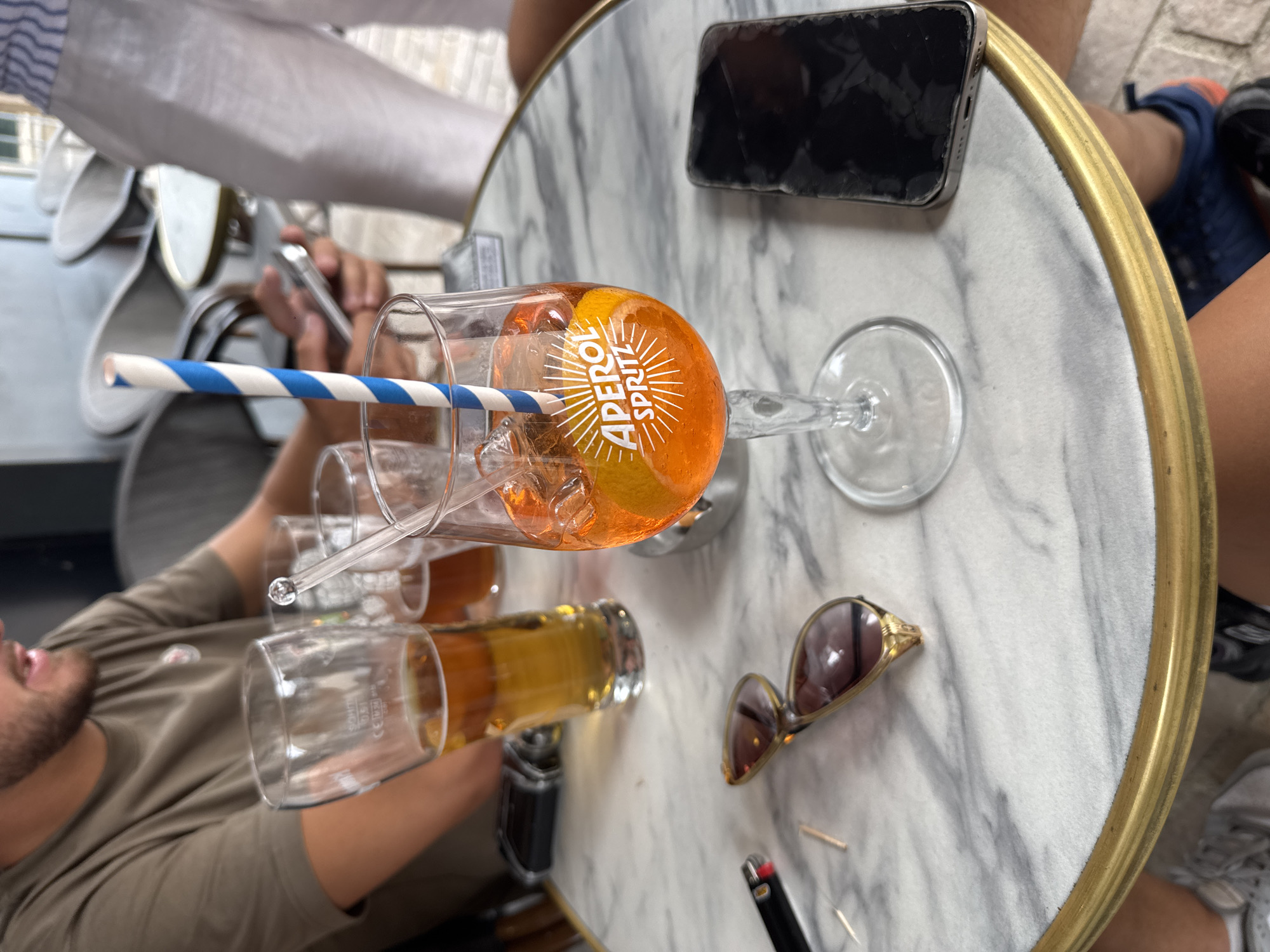 In Vienne, France, on a marble-topped table at an outdoor cafe, an Aperol Spritz sits prominently in a stemmed glass, garnished with an orange slice and a blue and white striped straw. Beside it, a glass of amber liquid, possibly beer, is also visible. Other objects on the table include a black lighter, a pair of sunglasses with brown lenses and gold frames, and a smartphone with a cracked screen. In the background, several people are seated at tables and chairs, suggesting a relaxed social setting. The lighting indicates it is daytime, likely an afternoon.