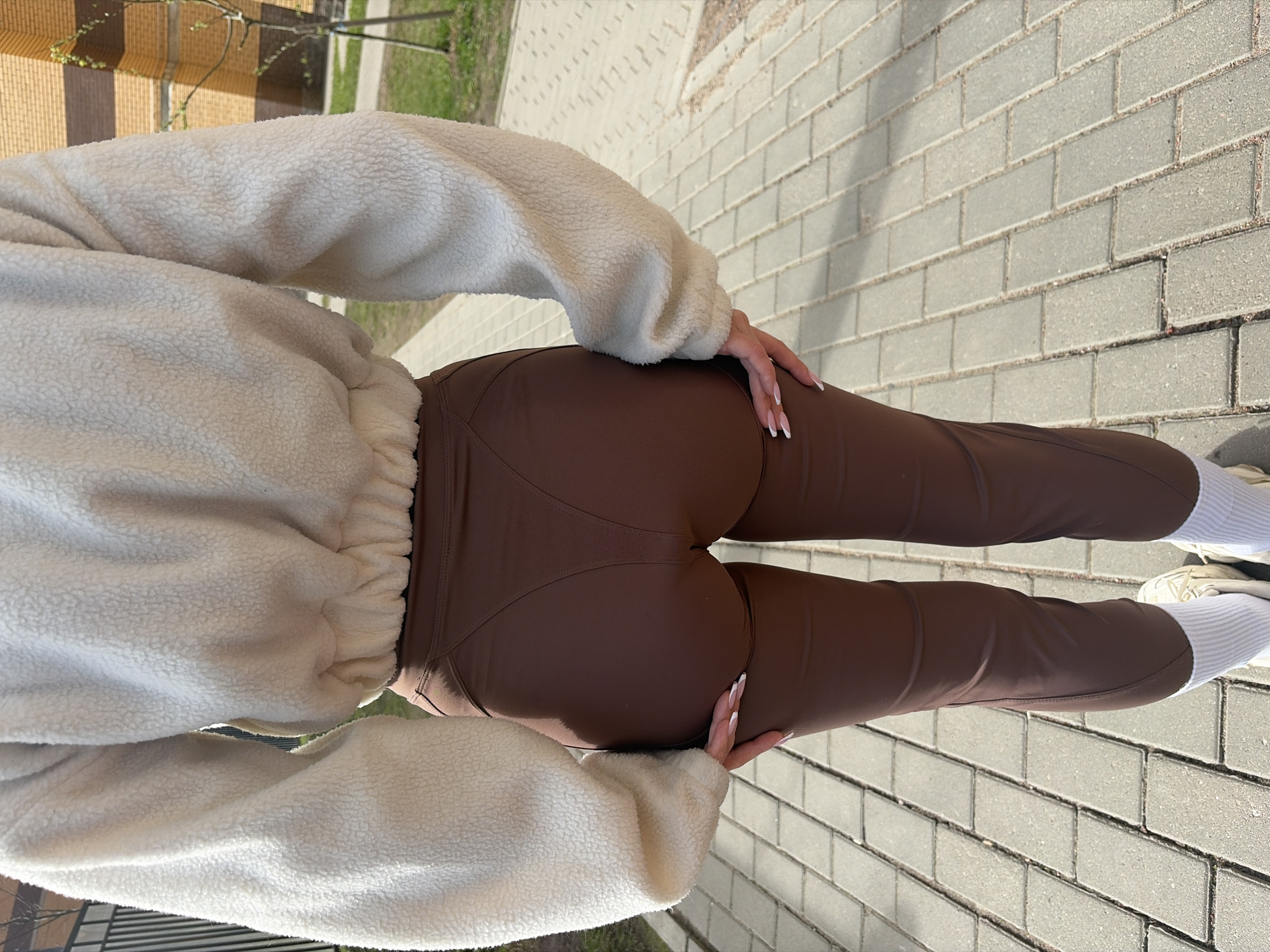 A woman wearing a cream-colored fleece sweatshirt and brown leggings is standing on a sidewalk made of gray rectangular pavers. She is seen from the rear view. Her hands are resting on her thighs. She is also wearing white socks and sneakers. There are trees and a building in the background. The scene is outdoors and appears to be in daylight. The specific location within Saint Petersburg, Russia, cannot be determined from the visual information provided.