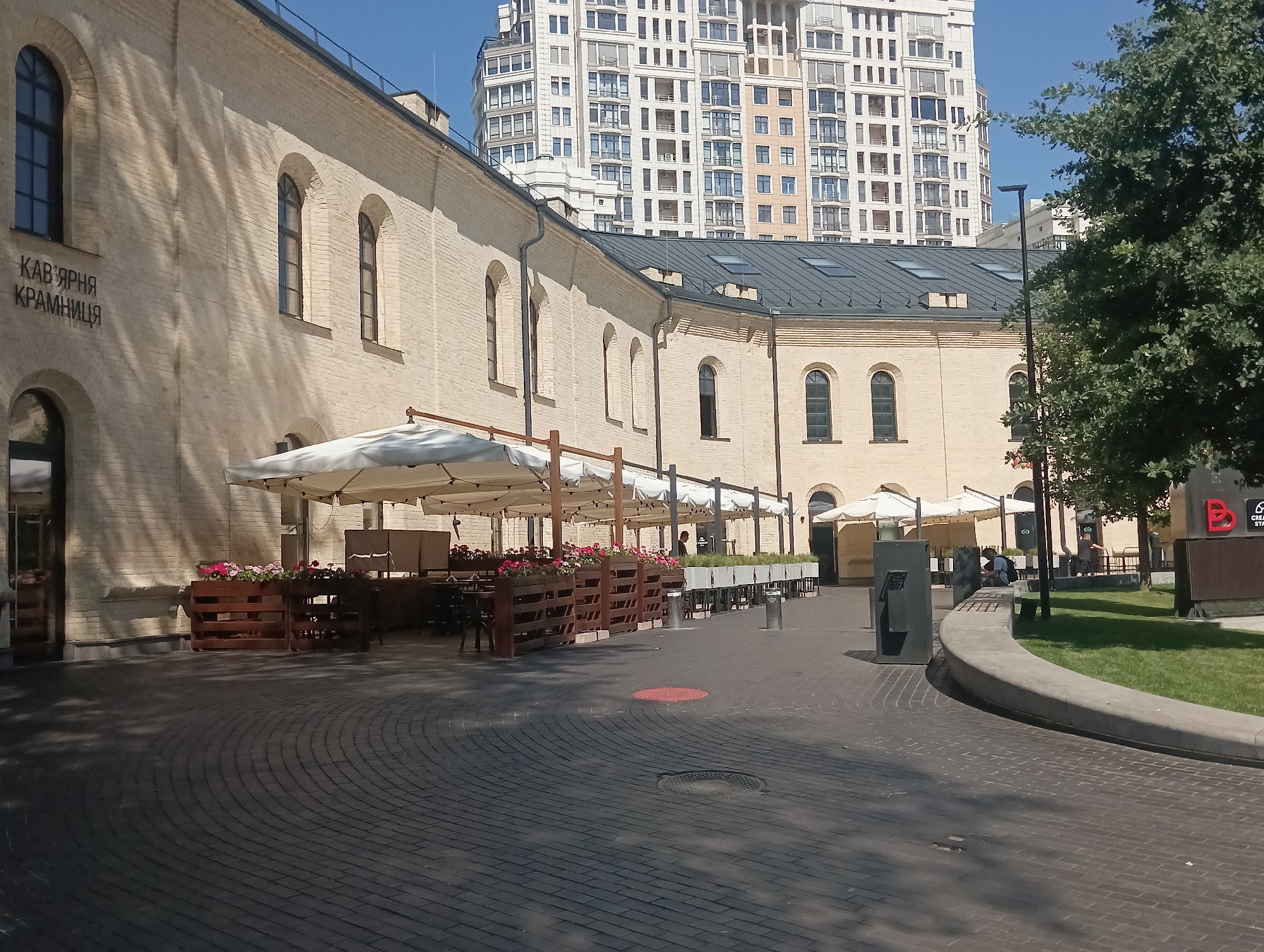 The image shows an outdoor seating area of a cafe, likely located in Kyiv, Ukraine. A long, pale brick building with arched windows forms the backdrop to the seating area, which is furnished with tables and chairs under white umbrellas. Flower boxes filled with pink flowers line the front of the cafe. In the background, a modern, tall apartment building stands against a clear blue sky. The ground is paved with dark bricks. There is a single person visible, sitting on a bench in the middle distance, looking down at something. The scene appears to be a bright, sunny day.