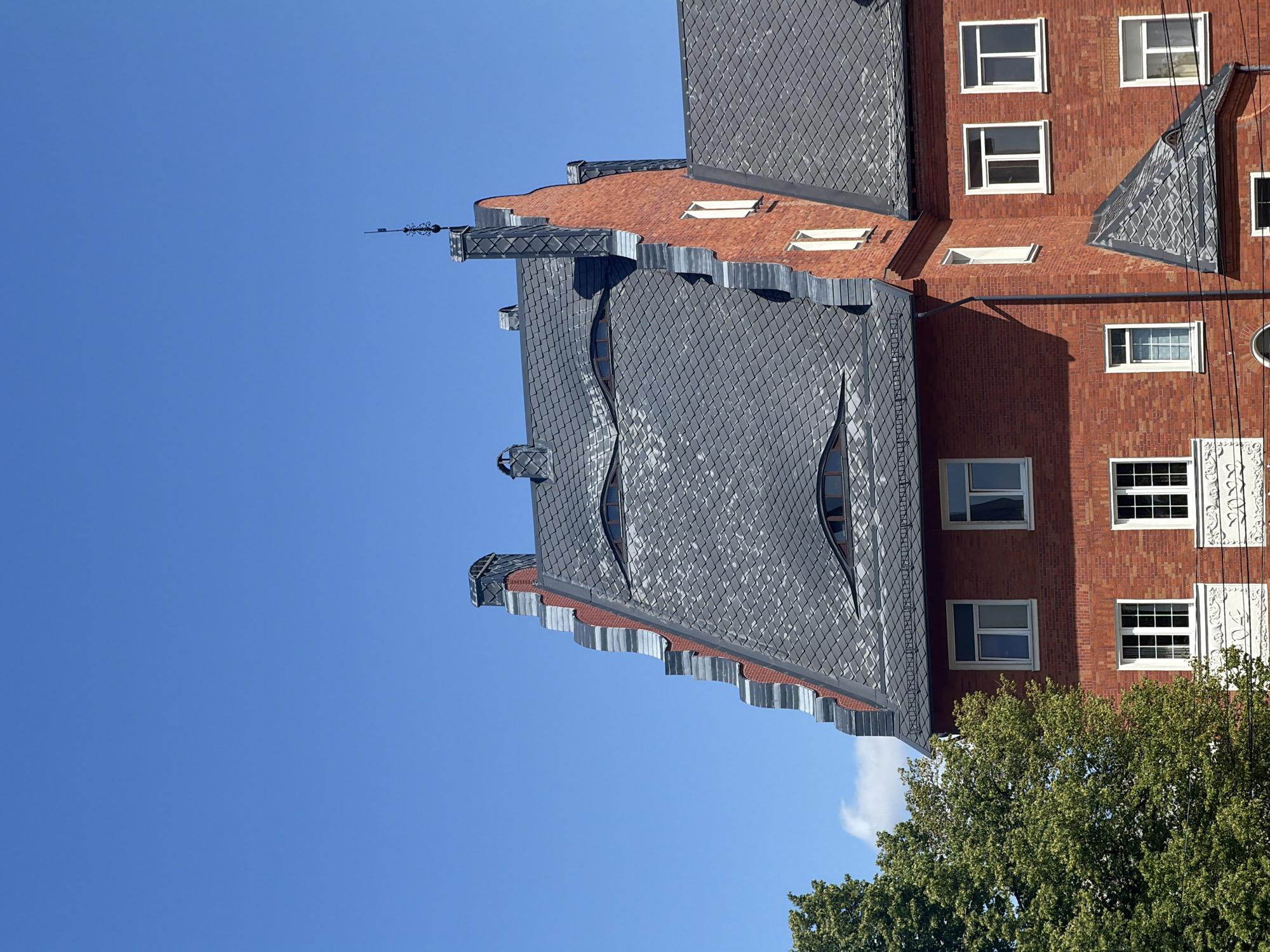 This media file depicts a close-up, slightly angled view of an apartment building in Kaliningrad, Russia. The building has a distinctive architectural style, featuring red brick walls and a dark gray, diamond-patterned tiled roof. The roof has several unusual, wavy gables and dormer windows with curved glass panes. The overall impression is one of a modern building with some unique, decorative elements.
The scene is set against a clear, bright blue sky, suggesting a sunny day with good weather. The sun casts distinct shadows on the building, indicating it is likely daytime, possibly late morning or early afternoon, given the angle of the light. The presence of lush green trees at the bottom right of the frame suggests a well-maintained urban environment.
There are no people visible in the image, and no specific activity or event is taking place. The focus is entirely on the building's architecture and its surroundings. Notable details include the intricate metalwork on the rooftop spire, the staggered pattern of the roof tiles, and the varied design of the windows. The red brickwork is also a prominent feature, showing a classic building material. The building appears to be residential, with multiple windows suggesting individual apartments.
There is no visible text in the image. The overall mood is one of architectural appreciation, highlighting the building's unique design against a serene natural backdrop.