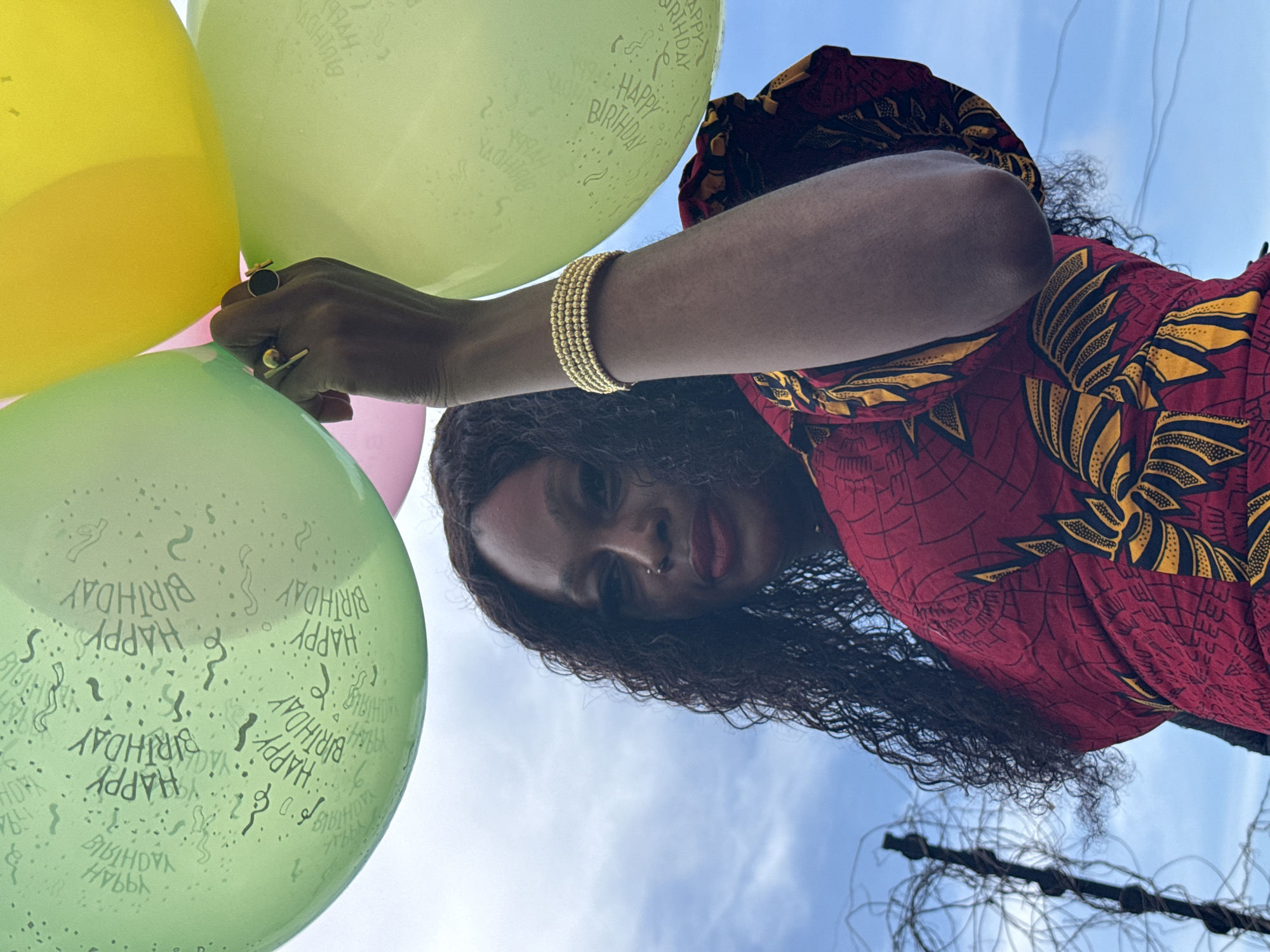 A low-angle photograph captures a person holding a cluster of colorful balloons against a bright, cloudy blue sky. The subject, a woman, has dark skin, long, dark curly hair, and is looking down towards the camera. She wears a vibrant red dress adorned with distinct yellow and black geometric patterns. Her accessories include a multi-strand gold bead bracelet on her left wrist and two rings on her left hand – one with a circular black stone and another with a gold bar design. She also has a small nose piercing.
The balloons are in shades of yellow, light green, and pink. The light green balloons prominently feature the text "HAPPY BIRTHDAY" printed in repeating patterns, indicating a celebratory occasion. The background consists of a clear, pale blue sky with scattered white clouds, suggesting a daytime outdoor setting. A dark, tangled structure, possibly wires or branches, is subtly visible in the bottom right corner of the frame. The scene evokes a festive and joyful atmosphere, likely a birthday celebration taking place outdoors in Igboefon, Nigeria.