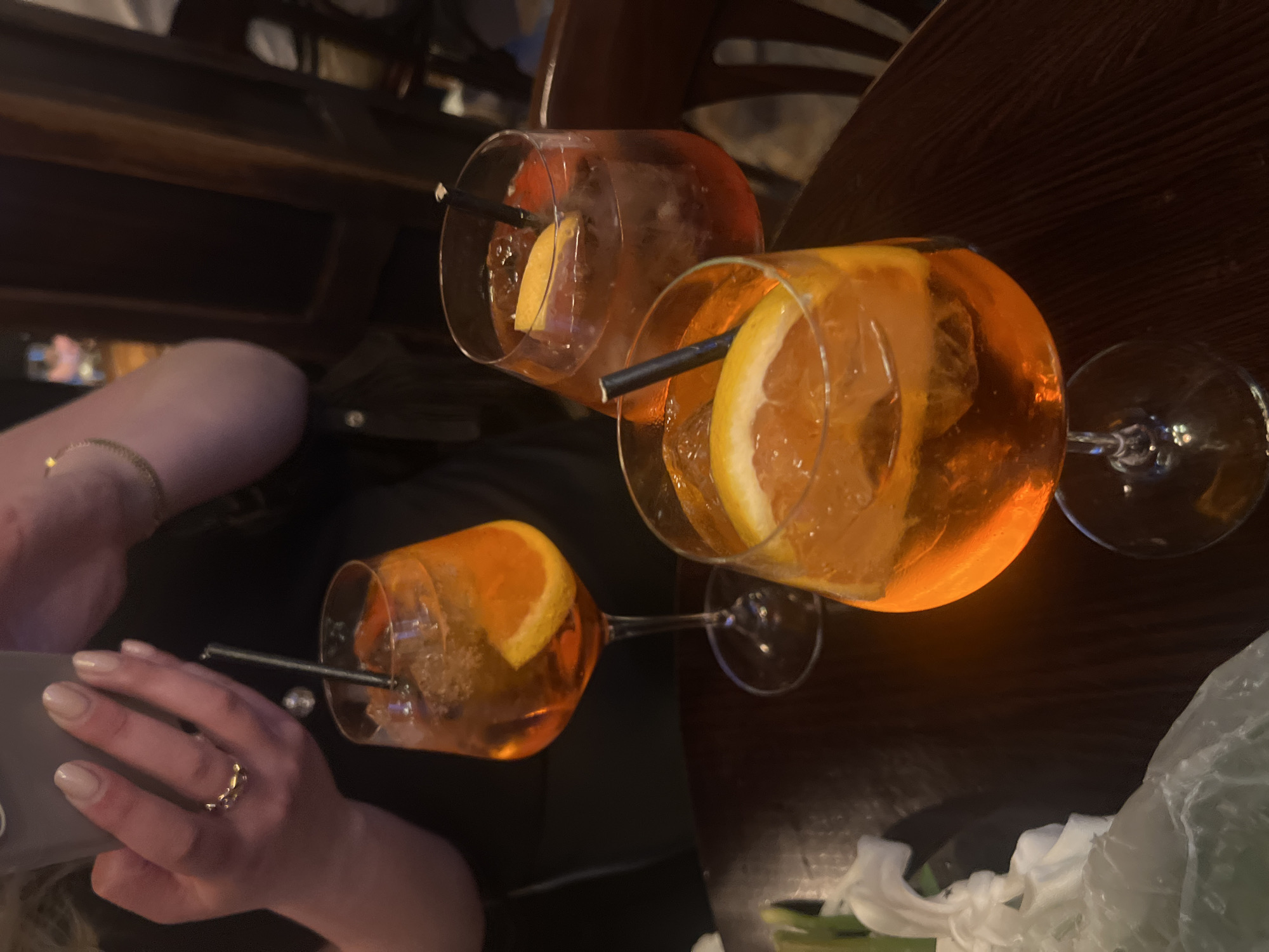 Three glasses of Aperol spritzes are on a dark brown table. Each glass has a slice of orange and a black straw. A person is holding a phone with their left hand near one of the glasses. The person is wearing a ring on their finger. Parts of another person are visible on the left and upper left of the image. The location is likely a bar or restaurant. The precise location in London is not identifiable based on the image.