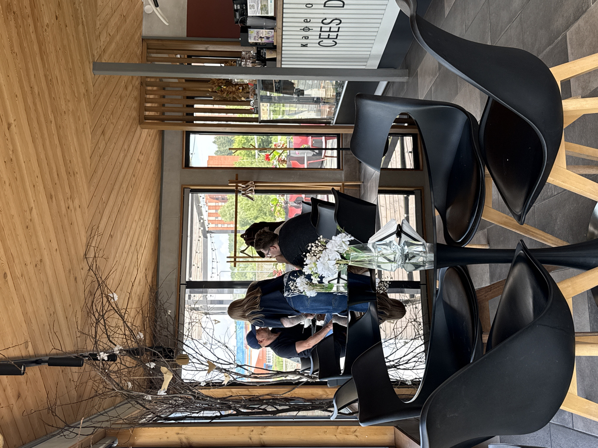 The image shows the interior of a cafe, likely in Votkinsk, Russia, with large windows offering a view of the outdoors. The cafe features a modern aesthetic with black tulip chairs around a round, reflective black table. The ceiling is made of light-colored wood planks. A decorative dry tree with small white paper birds hangs near the window, adding a whimsical touch.
Inside, a group of people are seated at tables. In the foreground, a black table with a glass vase of white flowers is prominent. In the background, near the window, a man wearing a black cap and a black polo shirt is seated, facing a woman who is standing and appears to be a server. The server, wearing a dark top and denim apron, is interacting with the man. Another person is visible at a table further in the background.
Beyond the windows, a glimpse of the urban environment is visible, with buildings and trees suggesting a city setting. The cafe's counter area is visible on the right side of the image, featuring a display case and signage that includes Russian text and the words "CEES D."