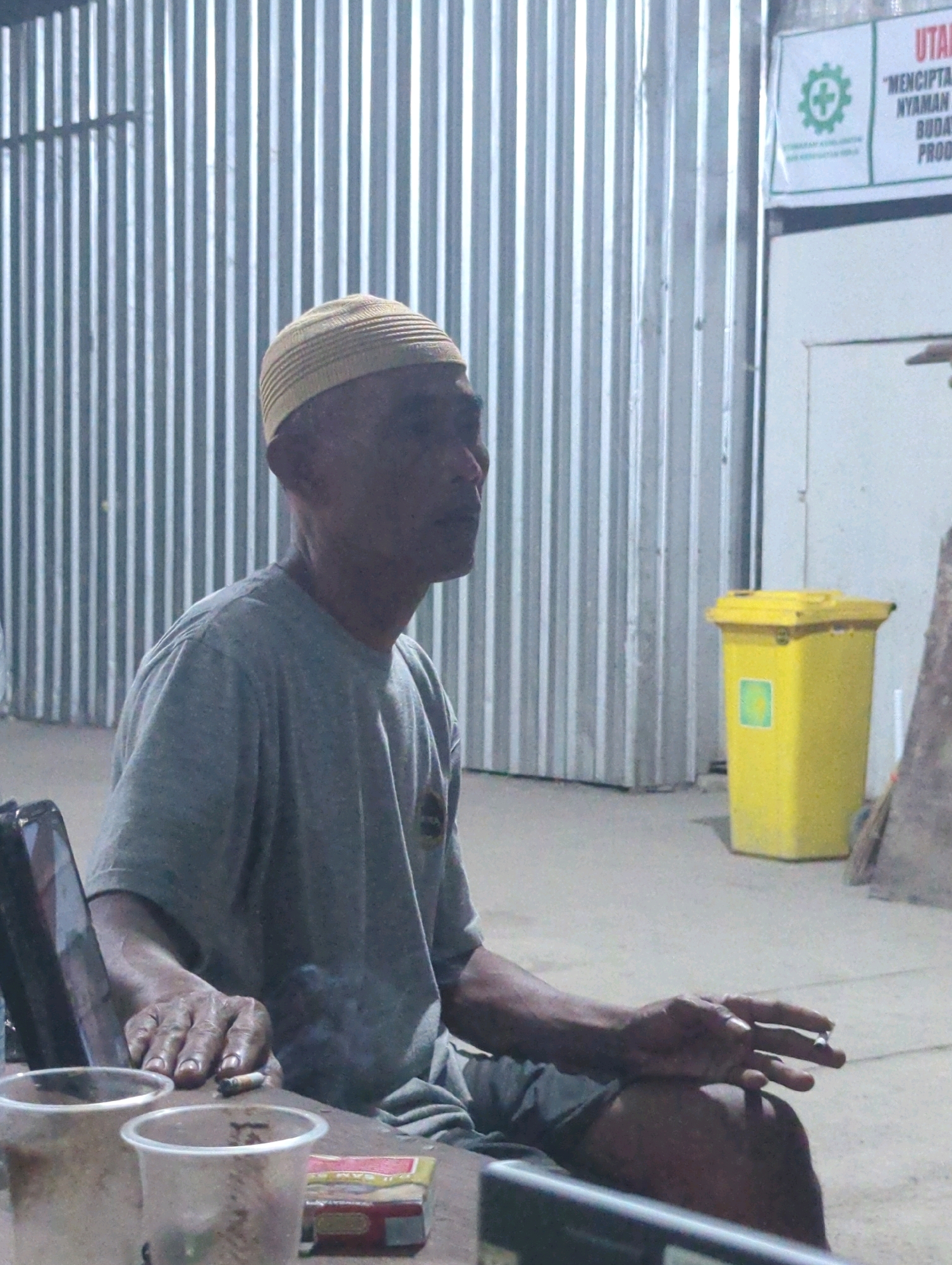 An elderly Indonesian man wearing a yellow kufi and a grey t-shirt sits outdoors in Batu Belaman, Indonesia, holding a cigarette in his right hand. He is seated at a wooden table with two disposable cups and a pack of cigarettes. Behind him, a corrugated metal wall forms the backdrop, and to his right, a bright yellow trash bin stands against a white wall. The lighting suggests it is dusk or night.