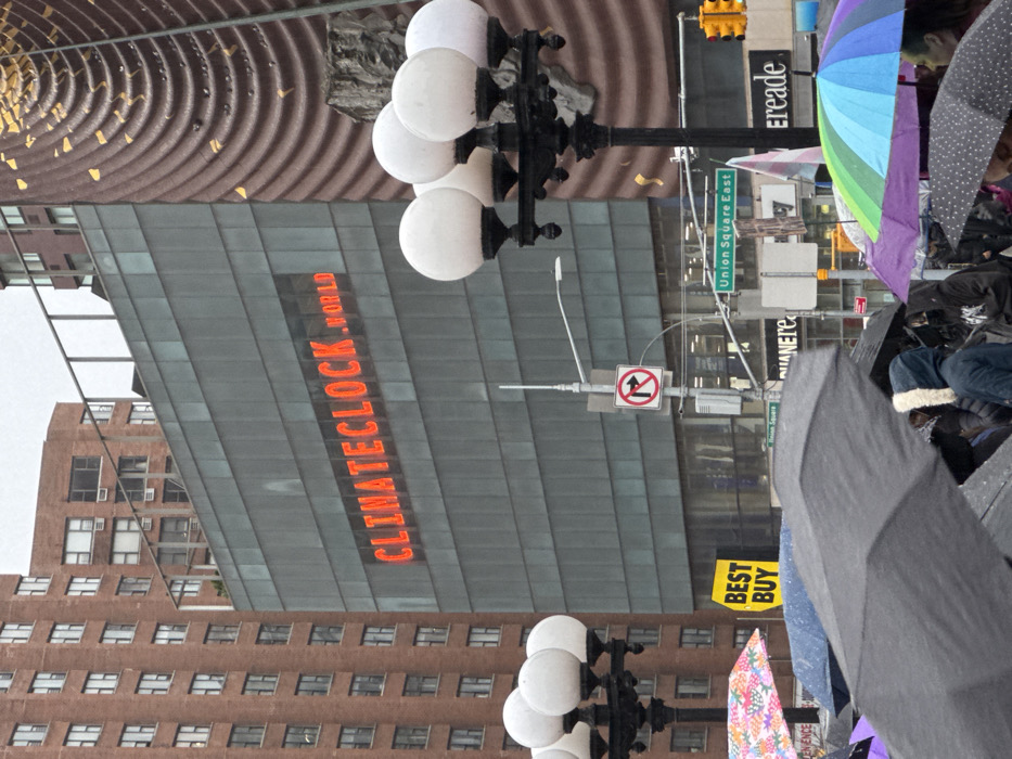 A street scene shows the "CLIMATE CLOCK by WORLD" signage on a building. Street signs for "Union Square East" are visible. A traffic light is present. Umbrellas, some colorful and some gray, are in the foreground. A Best Buy banner is on a sign. Street lights with multiple globes on poles appear in the foreground and middle ground. Buildings in the background display numerous windows.