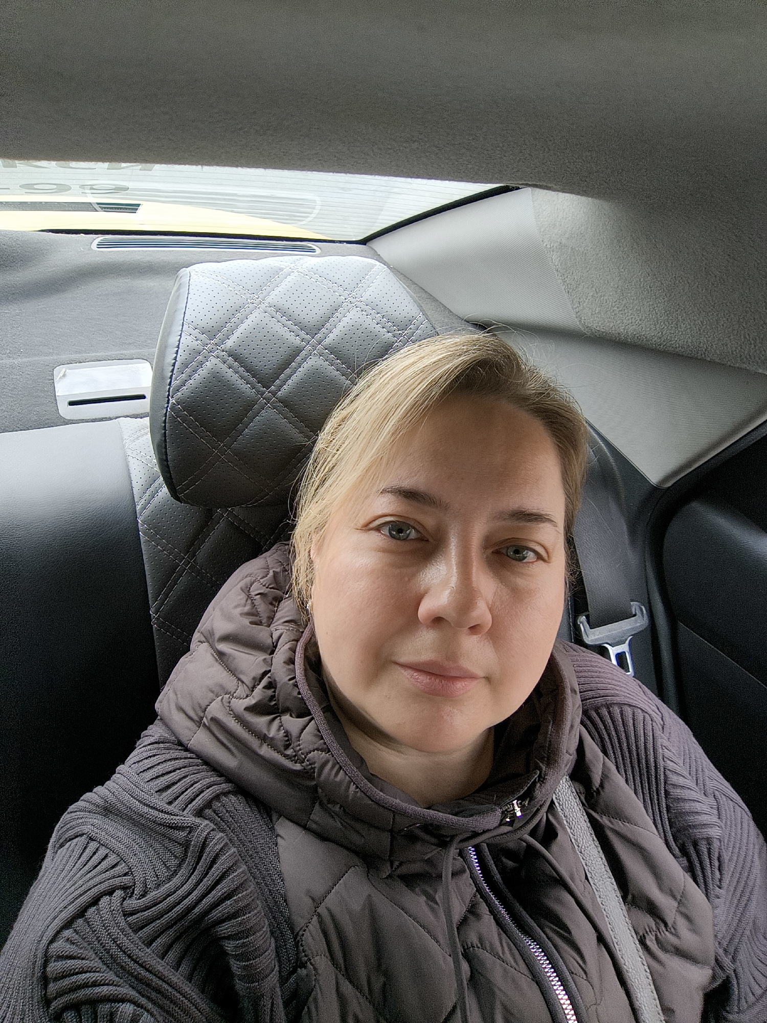 A woman is in the backseat of a car. She has fair skin, light hair, and is wearing a grey quilted jacket over a grey sweater. She appears to be wearing a seatbelt. The car has leather seats with a quilted pattern on the headrest. The location is most likely not Khimki, Russia based on the photo.