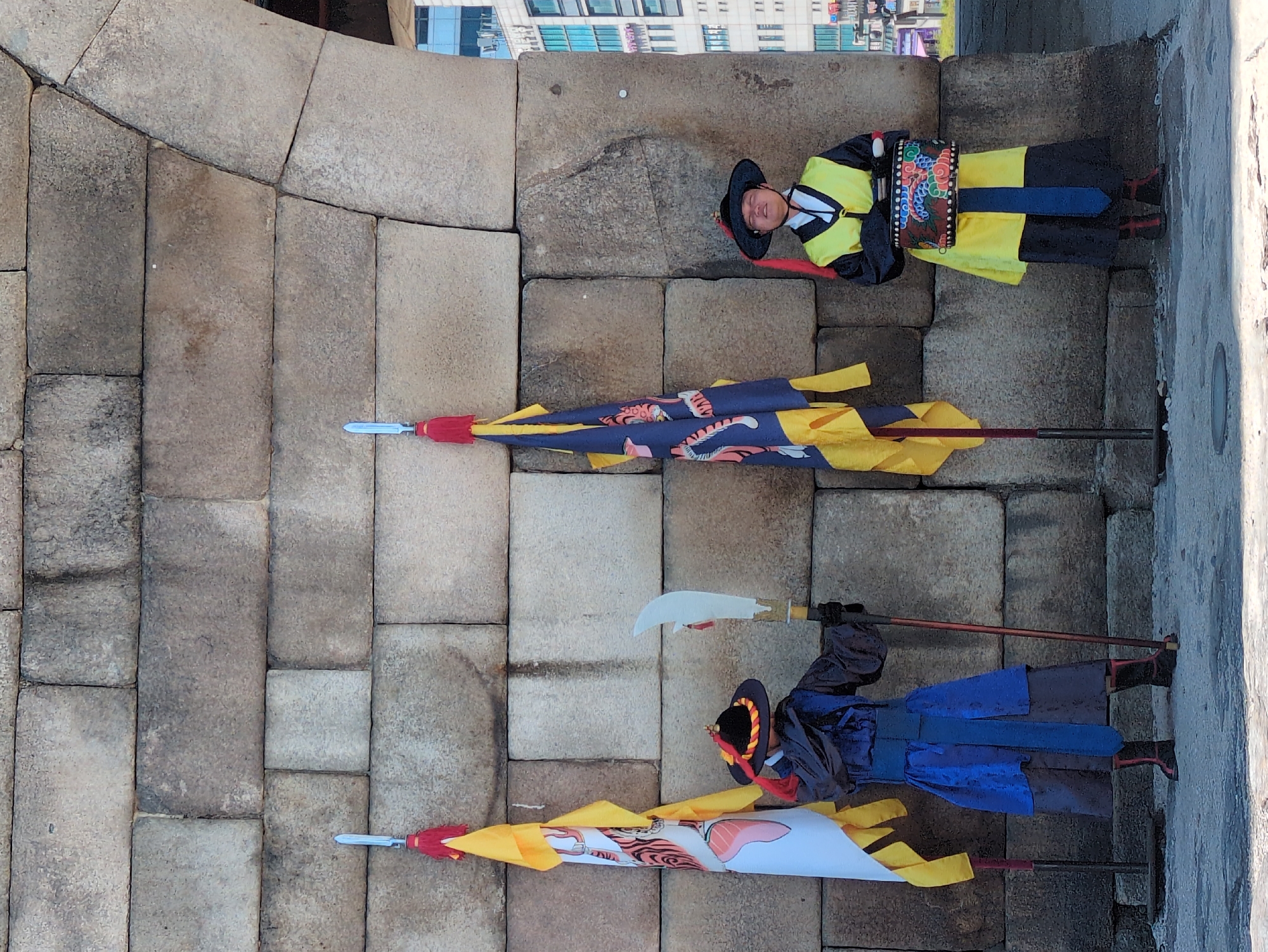 Captured from a high-angle perspective, the image showcases a scene in Seoul, South Korea.
A guard in traditional Korean attire, specifically a yellow robe with intricate designs, stands prominently. His hat is black and features a red string, and his belt is decorated. Adjacent to him, a spear with a blue and yellow banner imprinted with a tiger motif lies on the ground. Another spear with a white and yellow banner with a red and black design and a long, flat sword is positioned farther away.
The setting is a structure constructed from large, gray stone blocks, giving the impression of a historical building. Modern buildings can be seen in the background. The image's orientation suggests a portrait style, with objects appearing to be on the ground.