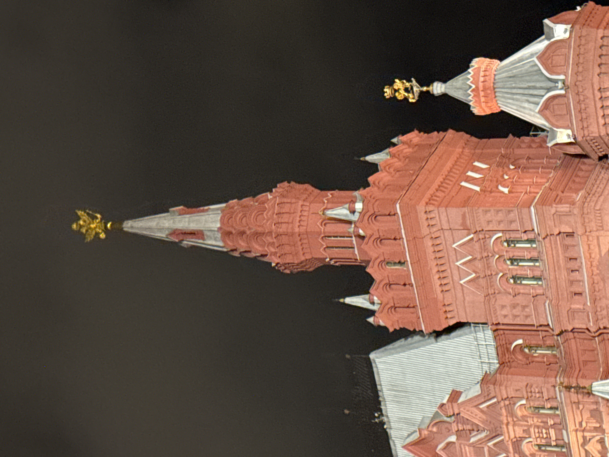 The image shows the illuminated towers of the Moscow Kremlin against a dark night sky. The Kremlin's red brick walls and architectural details are visible, topped with iconic spires and golden symbols. The scene is devoid of people, focusing solely on the architectural grandeur of the Kremlin at night.