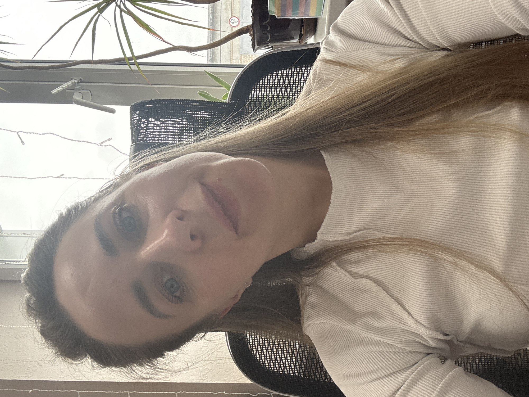 A woman with light brown hair and blue eyes is sitting in a black mesh office chair. She is wearing a white ribbed top. Behind her is a window with a plant and a view of a building. The location is potentially Chaikovsky, Russia.