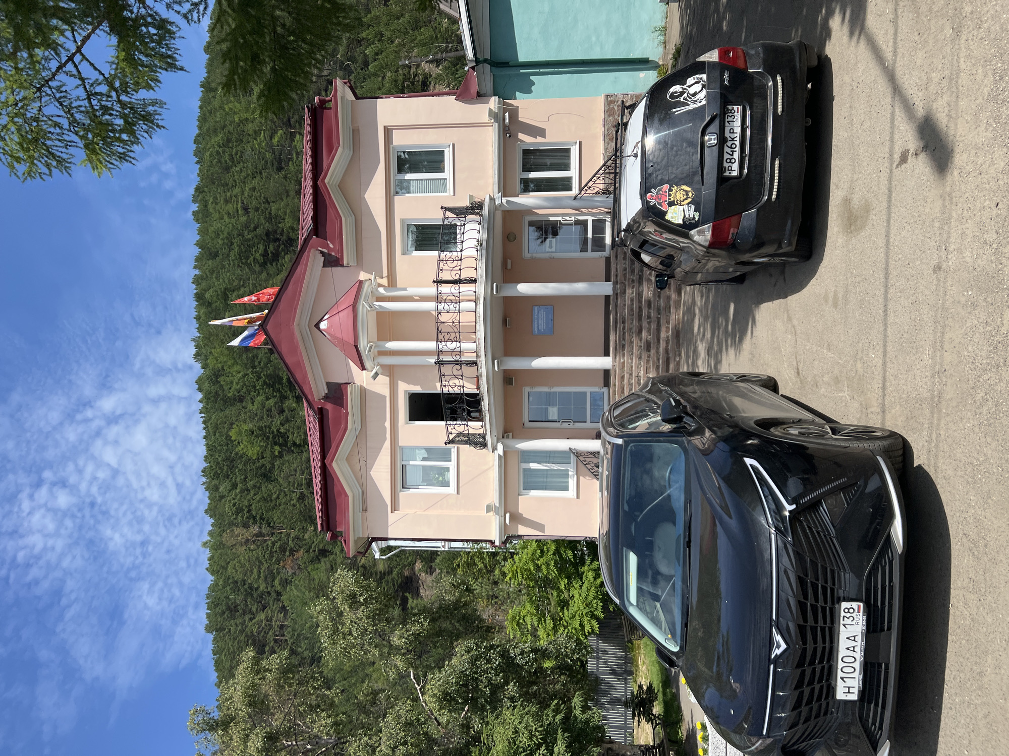 A two-story peach-colored building with a red roof stands on a slight incline, surrounded by a dense green forest. Three flags, including the Russian tricolor, fly from the roof. Two cars, a black sedan and a black hatchback, are parked in front of the building on a paved area. The sedan is in the foreground and is larger than the hatchback parked behind it. The hatchback is decorated with several stickers, including one of a person in a space suit and another of a lion's head. The sky above is a clear blue with scattered white clouds. The scene appears to be in Listvyanka, Russia, a village located on the southwestern shore of Lake Baikal.