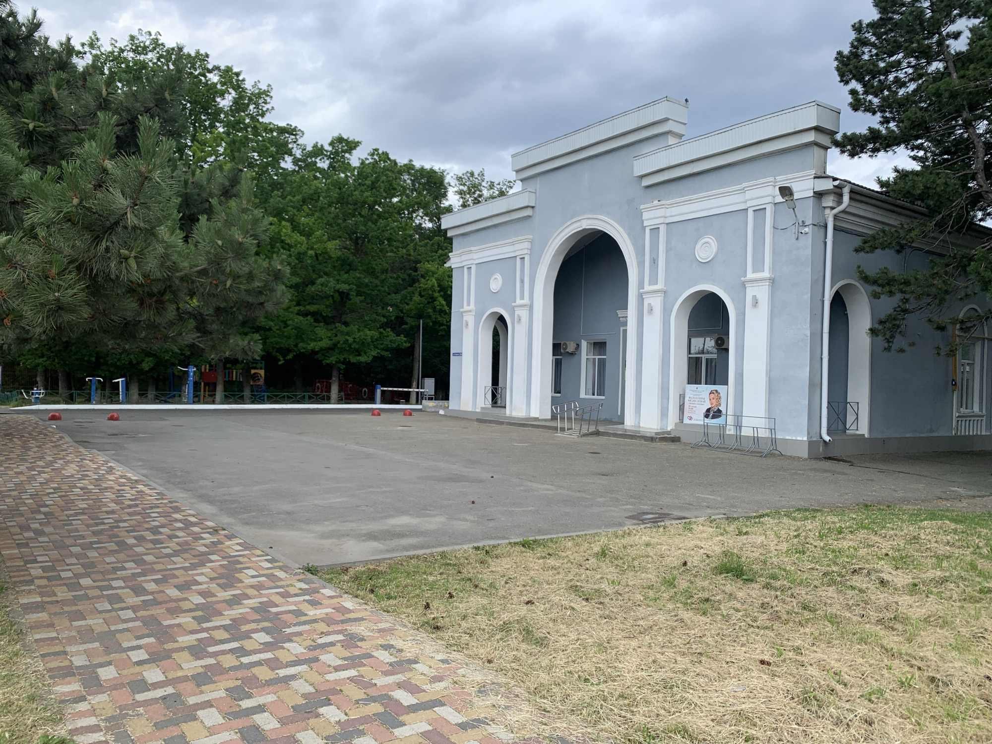 A pale blue, classical-style building with arched entrances and white trim is visible under a cloudy sky. In front of the building, there is a paved area with a sidewalk made of multi-colored rectangular bricks. Beyond the paved area, there is a grassy area with dry patches and scattered pinecones. In the background, there are lush green trees and a portion of a children's playground with colorful structures and red spherical bollards. A white barrier is also visible in the distance. A poster featuring a woman's portrait is displayed on the side of the building. This scene appears to be the entrance to a park or recreational area.