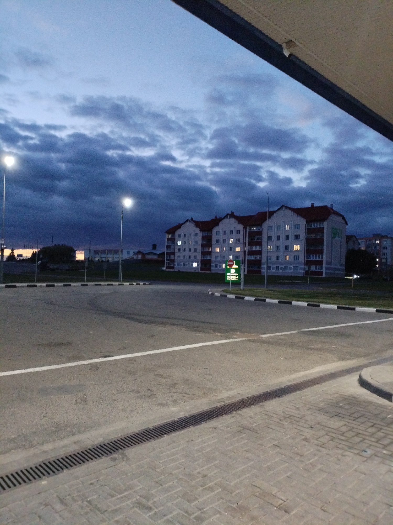The image depicts an outdoor scene at dusk, likely in Bialynichy, Belarus. A paved area with white road markings occupies the foreground, leading towards a row of buildings in the background. These buildings appear to be apartment complexes with visible lit windows. A street light is illuminated on the left side of the frame. In the mid-ground, there is a traffic signal with a green illuminated sign. The sky is overcast with dark, heavy clouds. A small section of a building's awning is visible in the upper right corner of the image. The overall atmosphere is dim and overcast, characteristic of evening or twilight.