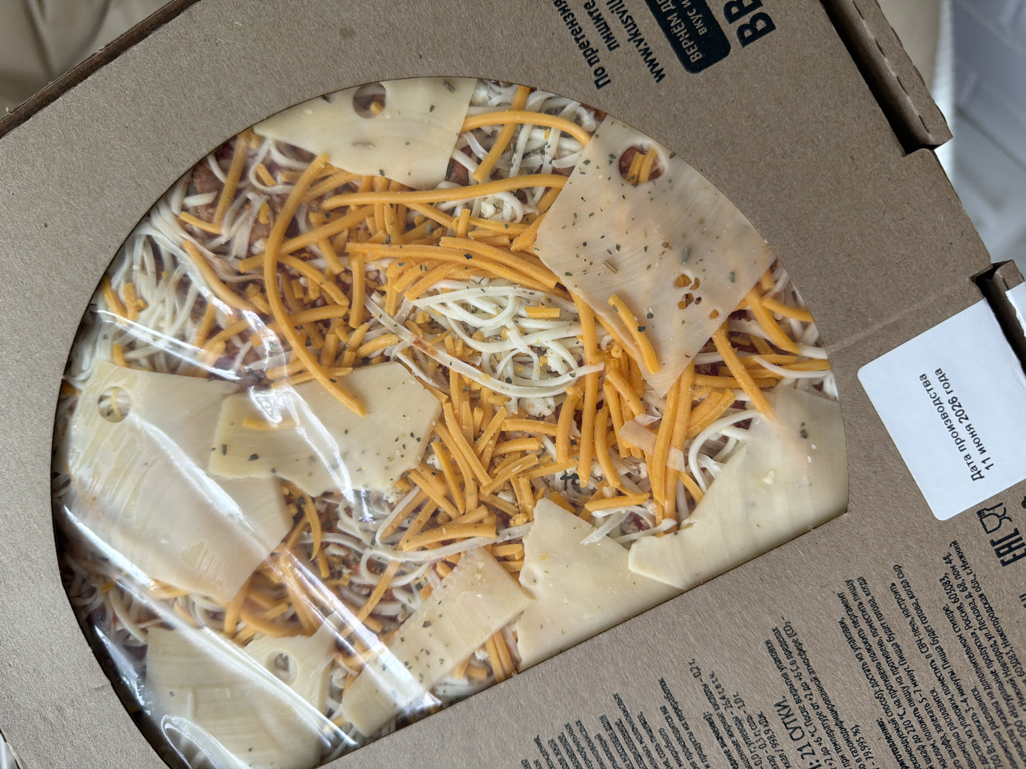 The image shows a raw, unbaked pizza in a cardboard box. The pizza is topped with tomato sauce, shredded white cheese, shredded orange cheese, and large slices of light yellow cheese. Herbs are sprinkled on top of the cheese. The pizza is partially covered with a clear plastic film. The cardboard box has some text printed on it, including "www.vkusvill.ru" and "ВЕРНЁМ ДЕНЬГИ ЗА ВКУС И КАЧЕСТВО". A white sticker with text in Russian is also visible, stating "Дата производства 11 июня 2026 года" (Production date June 11, 2026). There is no indication of the city of Semiluki, Russia in the image.