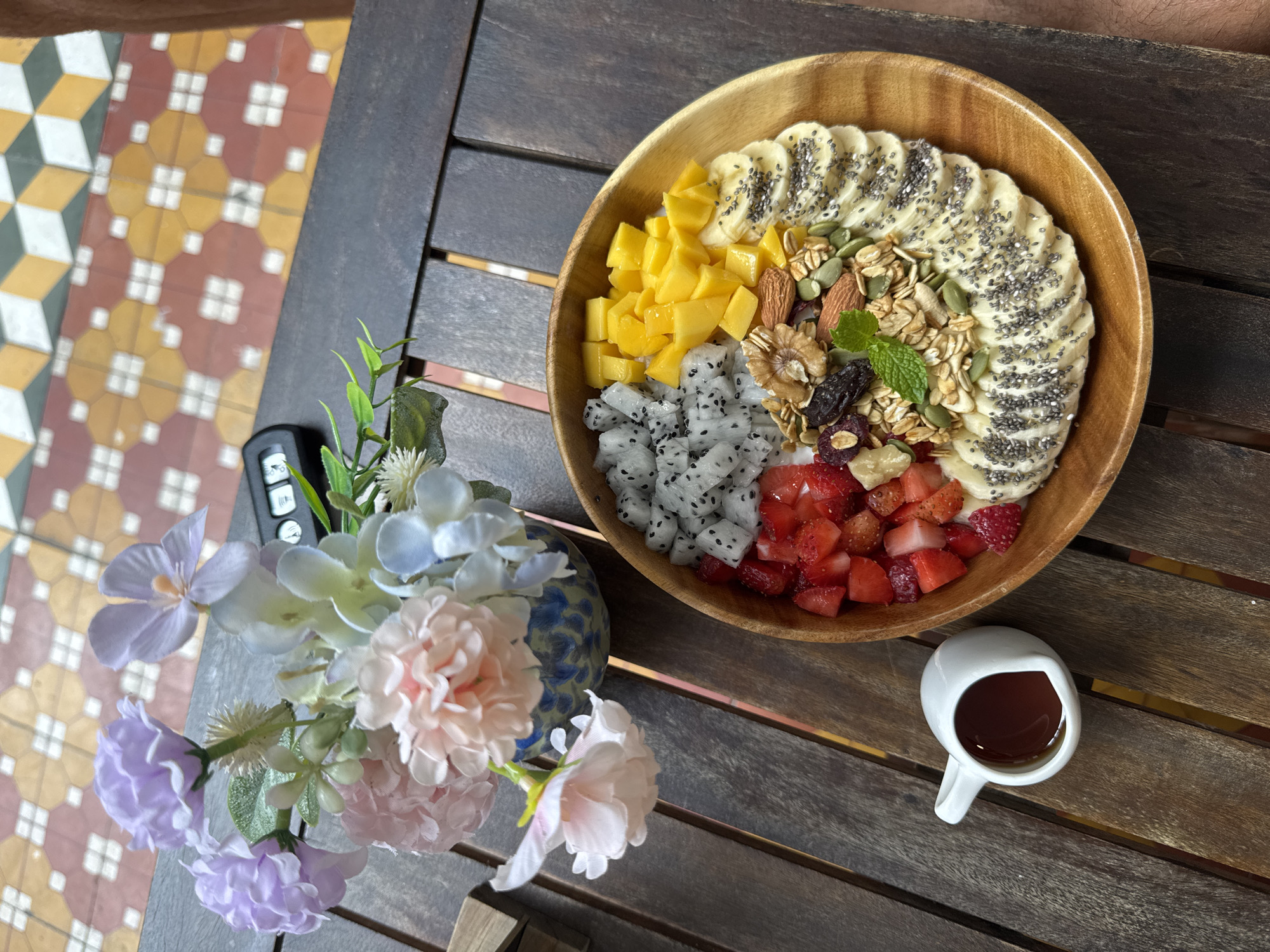A top-down shot depicts a wooden bowl of a smoothie bowl with assorted toppings, placed on a dark, stained wood table. To the left of the bowl is a small vase of pastel-colored flowers, and below the bowl is a small white mug filled with a dark liquid. In the bowl, clockwise from the top, are sliced bananas topped with chia seeds, granola with nuts, diced strawberries, diced dragon fruit, and diced mango. A car key is near the flowers. The floor tiles in the background suggest an interior space in Đà Nẵng, Vietnam.