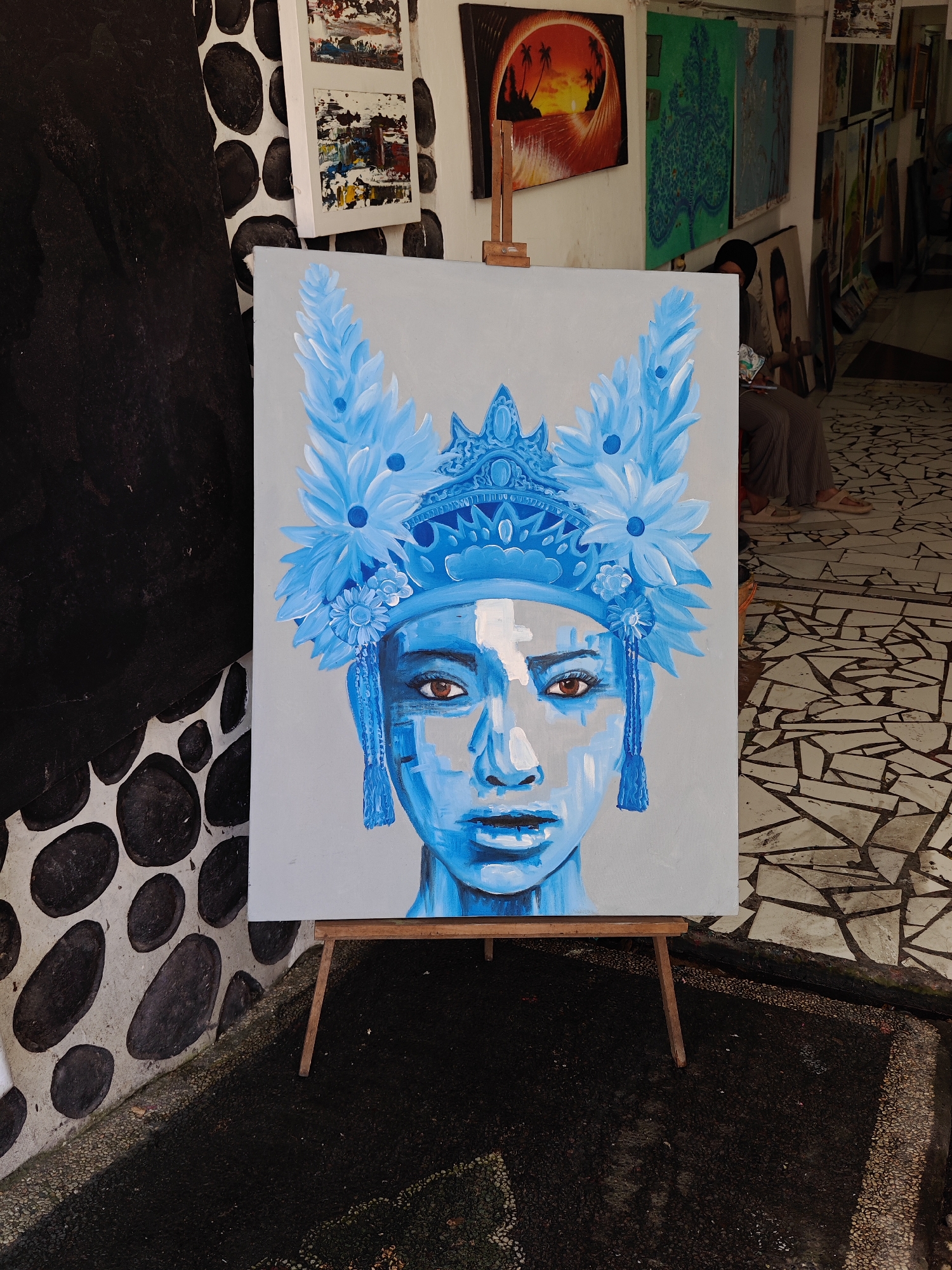 The scene is an outdoor art gallery in Denpasar, Indonesia. A large, framed painting depicting a woman with a blue complexion and headdress is positioned on a wooden easel. The painting is the central focus of the image. A person sits in the background, towards the right of the frame, with their face obscured. The floor is patterned with irregular white tiles. To the left, a wall is decorated with painted black ovals. Several other paintings hang on the wall behind the central artwork, including a piece depicting a sunset. The overall environment suggests a casual, art-focused setting in an urban environment.