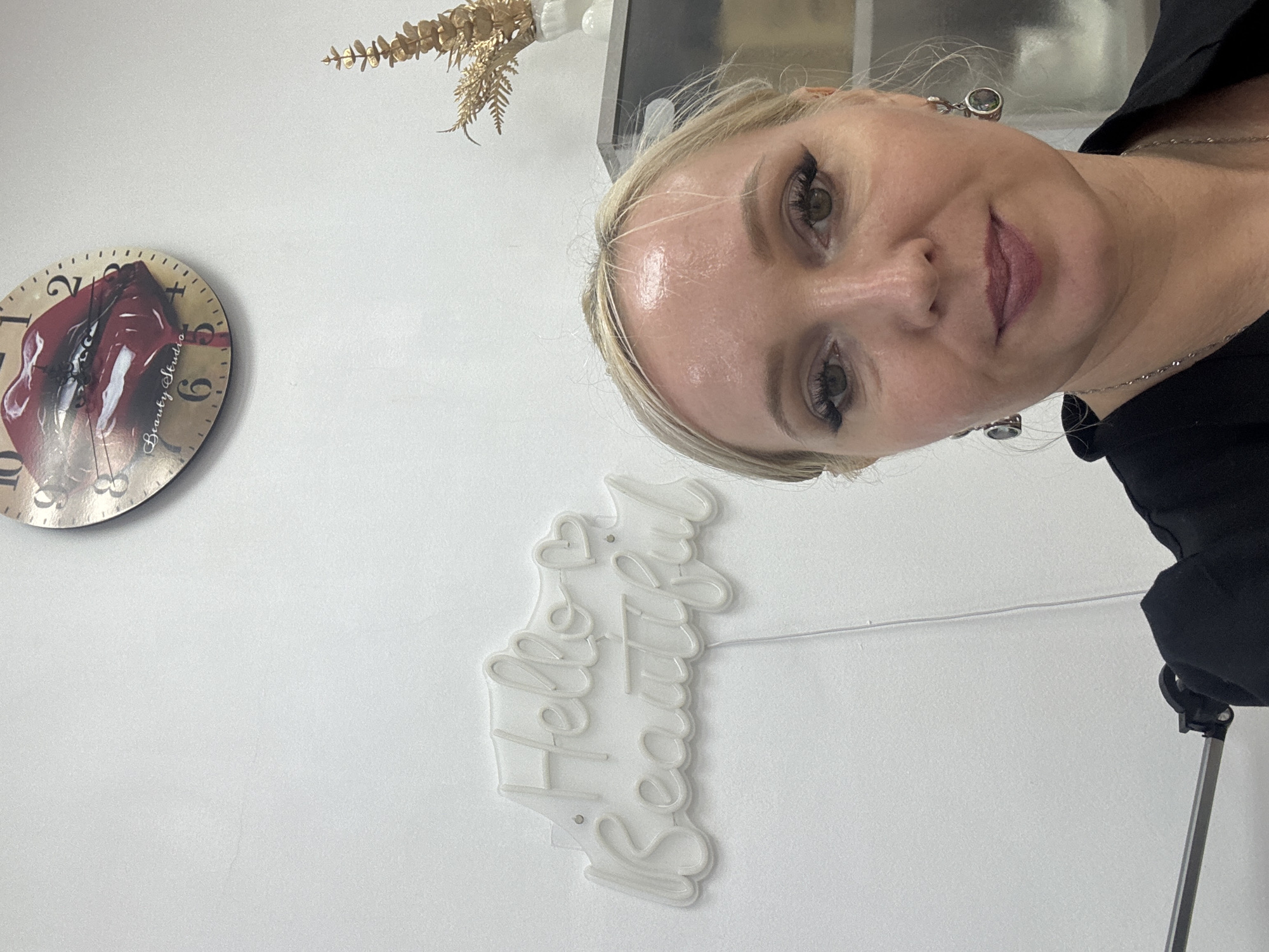 A blonde woman with makeup is taking a selfie in front of a white wall. A neon sign that reads "Hello Beautiful" is on the wall to her left, and a clock with a picture of red lips is on the wall above it. A golden decorative plant is on a shelf behind her to the right. The woman is wearing a black top and silver necklace. The scene appears to be in a beauty studio in Komsomolsk-on-Amur, Russia.