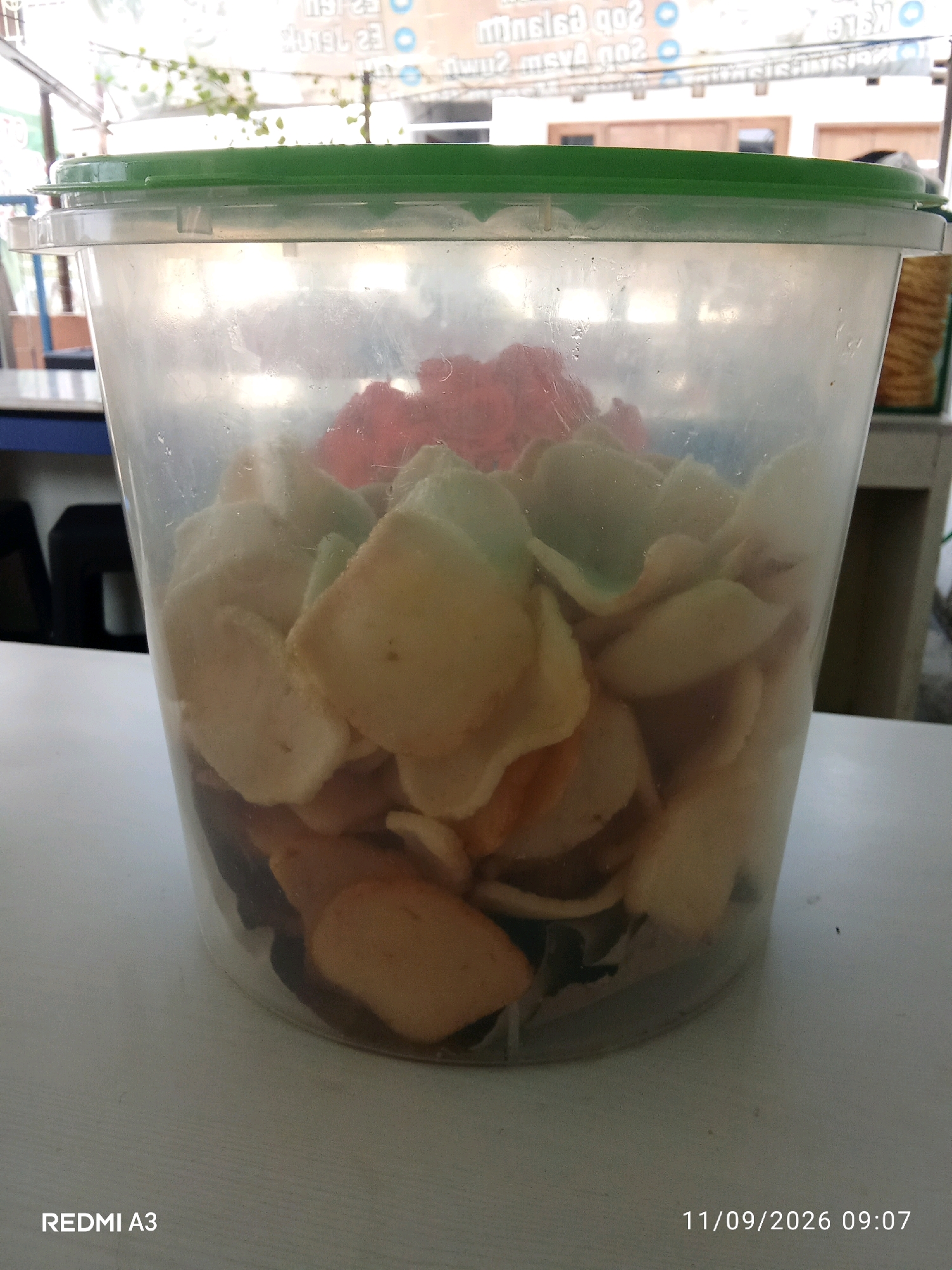 A clear plastic tub with a green lid is filled with light-colored crackers and some reddish items. The tub is placed on a light-colored, textured surface, likely a table. In the background, there is a blurred view of an indoor environment with a sign that has Indonesian text. The bottom right corner of the image displays the text "11/09/2026 09:07" and "REDMI A3". The scene is well-lit, suggesting it was taken during the day.
