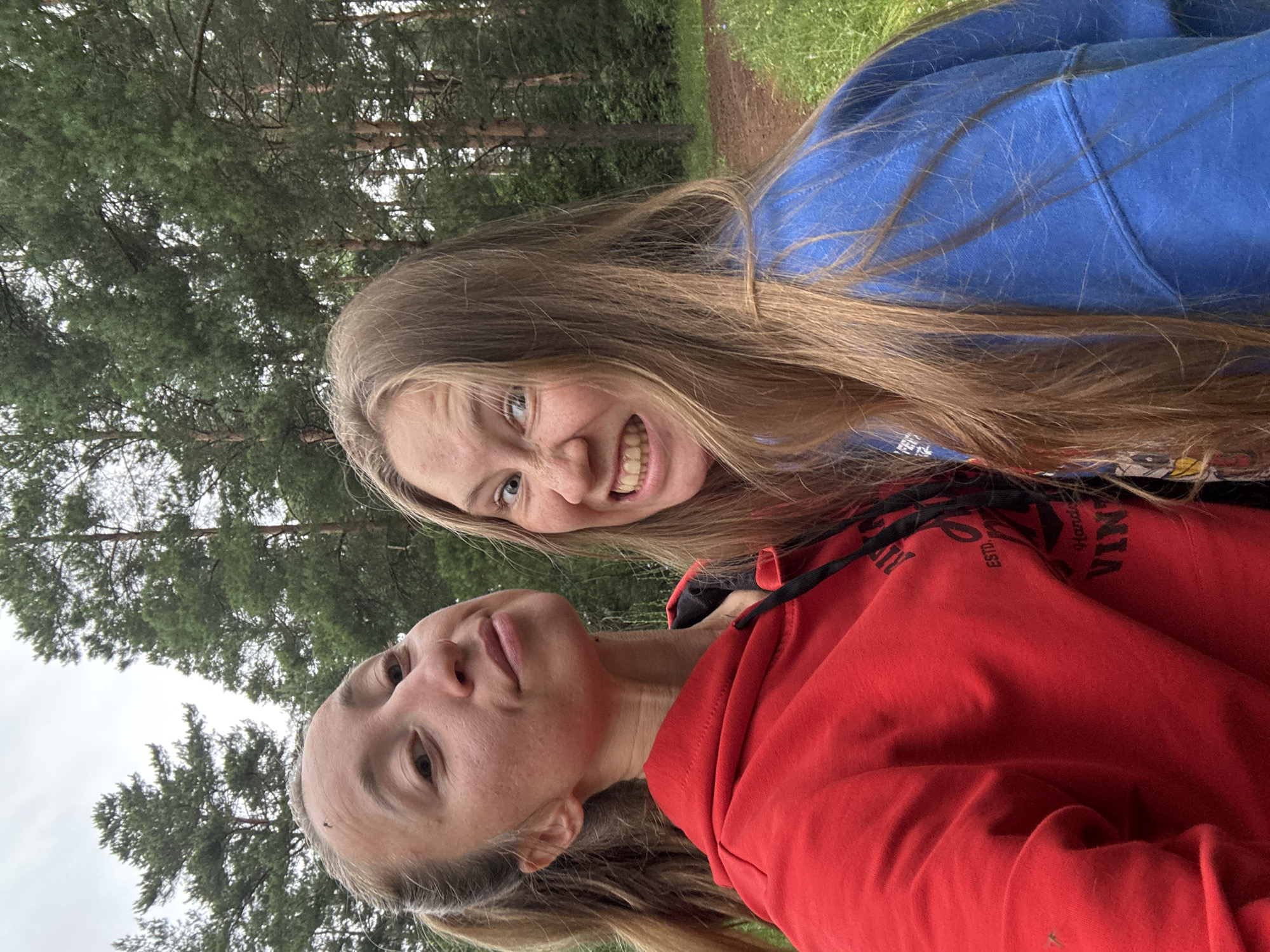 Two young women are posing for a selfie in an outdoor, wooded setting, likely in Прикамский, Russia. The woman in the foreground is wearing a bright red hoodie and is looking directly at the camera with a slightly playful expression. Her head is tilted, and her eyes are wide open. Her hair, which is long and light brown, is falling around her face and shoulders.
The second woman is positioned slightly behind and to the side of the first. She is wearing a blue hoodie and is looking towards the camera with a wide, toothy grin. Her long, light brown hair is also visible, framing her face.
The background consists of tall pine trees and green foliage, suggesting a forest or park area. A dirt path is visible in the upper right portion of the image. The lighting suggests it is daytime, and the sky appears overcast, indicating a cloudy day. The overall mood of the image is cheerful and casual, as the two women appear to be enjoying themselves. On the red hoodie of the woman in the foreground, partial text is visible, including "ESTD." and "VIN."