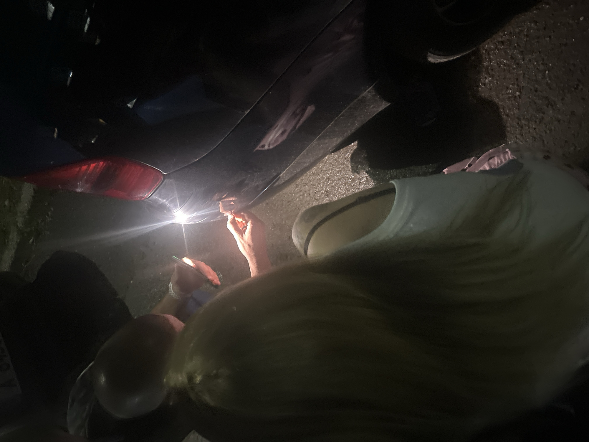 The image depicts a woman repairing a dark-colored car, possibly at night, in an unidentifiable location, with no evidence linking it to the city of Burgas, Bulgaria. The woman is using a light source, potentially a flashlight or mobile phone, to illuminate the damaged area on the car's side panel near the rear taillight. Her left hand holds a thin object that appears to be inserted into the damage. The scene's lighting is poor, and the environment seems to be a paved surface.